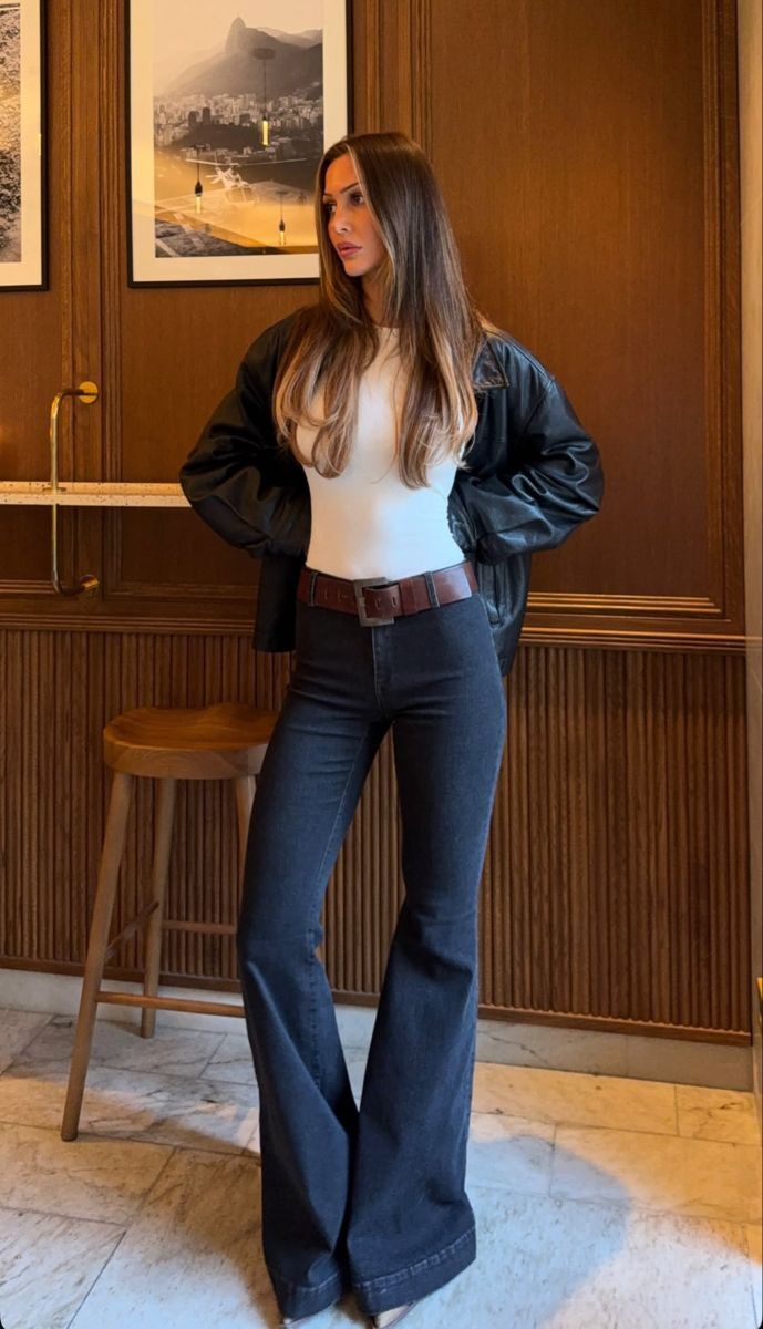 Mulher jovem, cabelos castanhos longos, veste blusa branca, jaqueta preta de couro, cal&ccedil;a jeans escura boca de sino e cinto marrom. Ela posa em p&eacute;, olhando para a direita, em um ambiente com pain&eacute;is de madeira e quadros na parede
