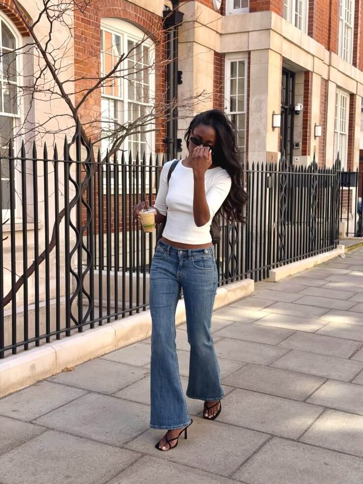 Mulher negra de cabelos longos e escuros, &oacute;culos de sol, blusa branca justa e cal&ccedil;a jeans boca de sino, caminhando em uma cal&ccedil;ada de paralelep&iacute;pedos. Ela segura um copo de caf&eacute; gelado na m&atilde;o esquerda e a m&atilde;o direita est&aacute; perto do rosto. Ao fundo, um pr&eacute;dio de tijolos e um port&atilde;o de ferro preto com detalhes ornamentados. Uma &aacute;rvore sem folhas est&aacute; &agrave; esquerda