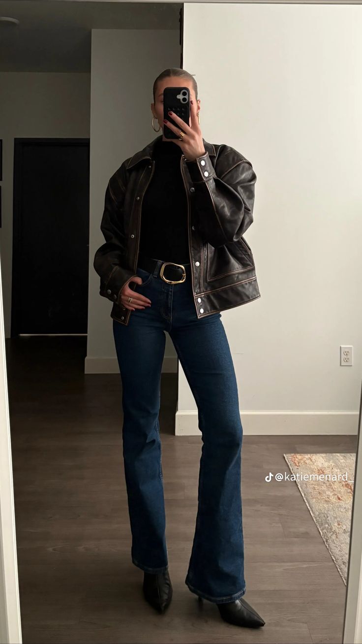 Mulher de p&eacute;, sem mostrar o rosto, tirando uma selfie no espelho. Ela veste jaqueta de couro marrom, blusa preta de gola alta, cal&ccedil;a jeans escura e cinto preto com fivela dourada. Usa brincos de argola e an&eacute;is dourados. O cabelo est&aacute; preso e as unhas pintadas de vermelho. O fundo &eacute; um ambiente interno com paredes claras e piso de madeira. O nome de usu&aacute;rio @katiemenard aparece no canto inferior direito