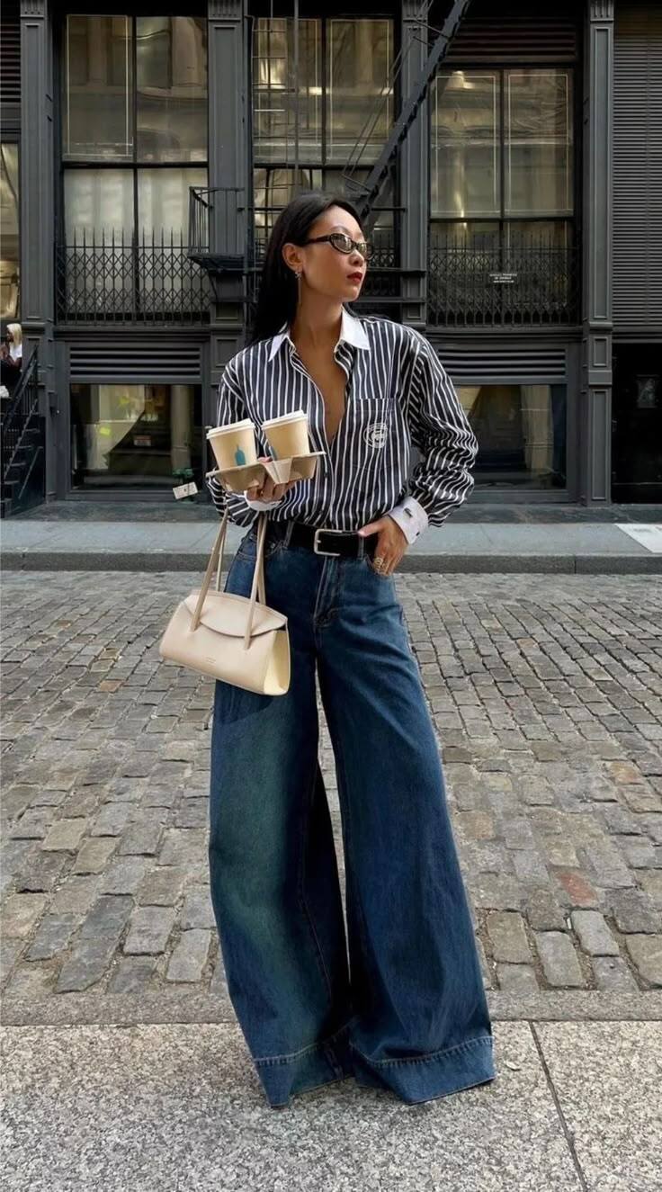 Mulher asi&aacute;tica com &oacute;culos, camisa listrada azul e branca, cal&ccedil;a jeans larga e bolsa creme, segurando dois copos de caf&eacute; em um suporte, em uma rua de paralelep&iacute;pedos com pr&eacute;dios escuros ao fundo