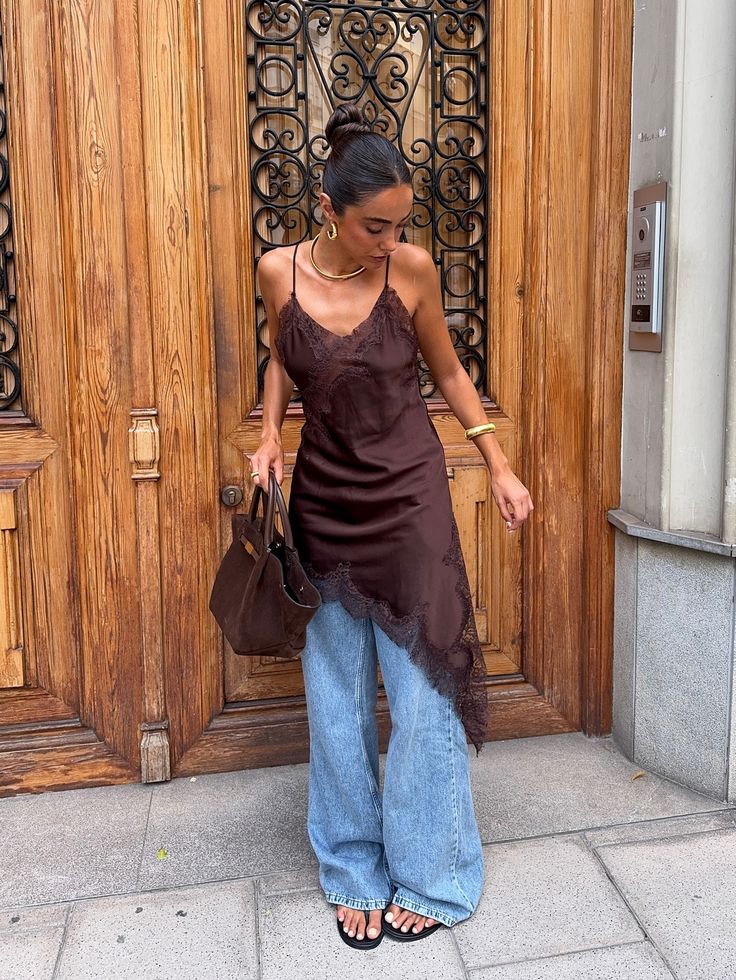 Mulher com coque alto, usando blusa de seda marrom com renda, cal&ccedil;a jeans larga, chinelos pretos e bolsa marrom, em frente a uma porta de madeira ornamentada