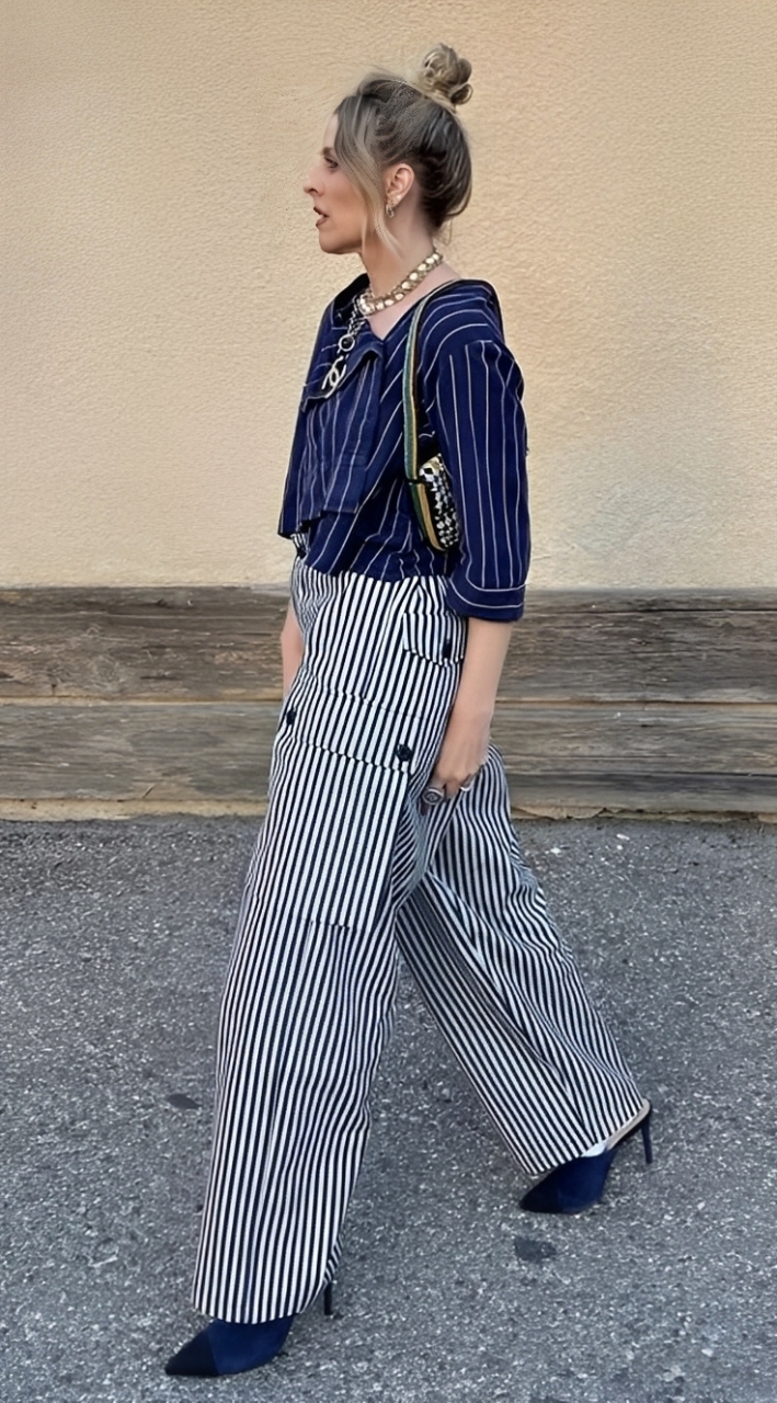 Mulher loira de coque, perfil esquerdo, usando camisa azul marinho listrada e cal&ccedil;a pantalona listrada em preto e branco, com bot&otilde;es pretos na lateral. Ela usa colar dourado, brincos pequenos e uma bolsa pequena de al&ccedil;a colorida, com detalhes em preto e branco. Cal&ccedil;a sapatos de salto alto azuis com bico preto, caminhando sobre asfalto cinza, com parede bege e faixa de madeira ao fundo