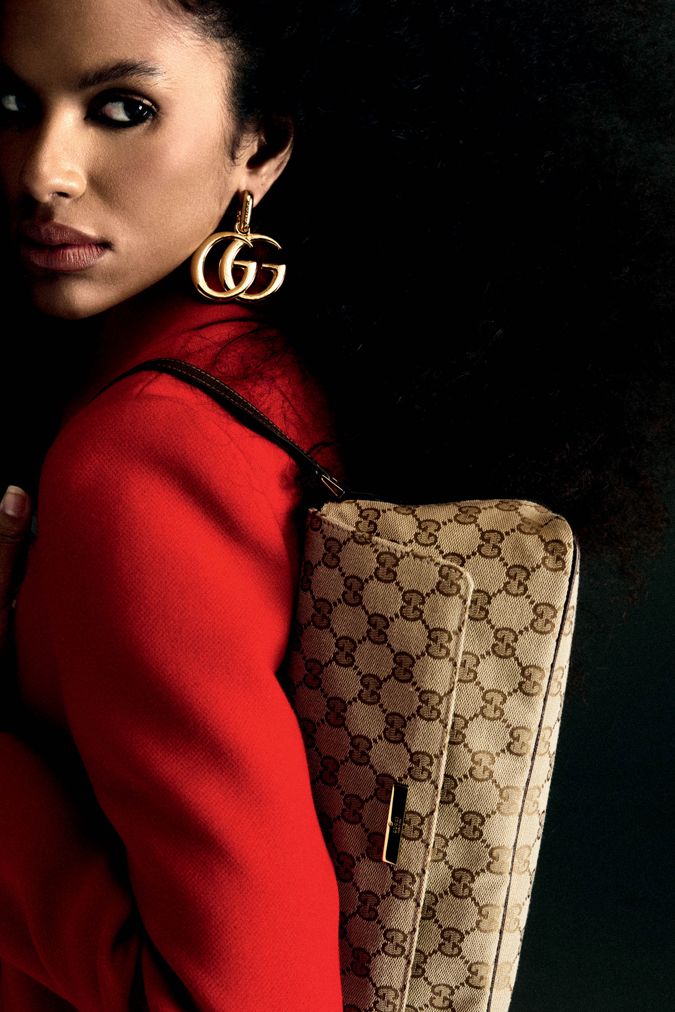 Mulher negra com cabelo crespo escuro, vestindo blazer vermelho, brinco dourado com o logo GG da Gucci e bolsa de ombro bege com monograma da marca