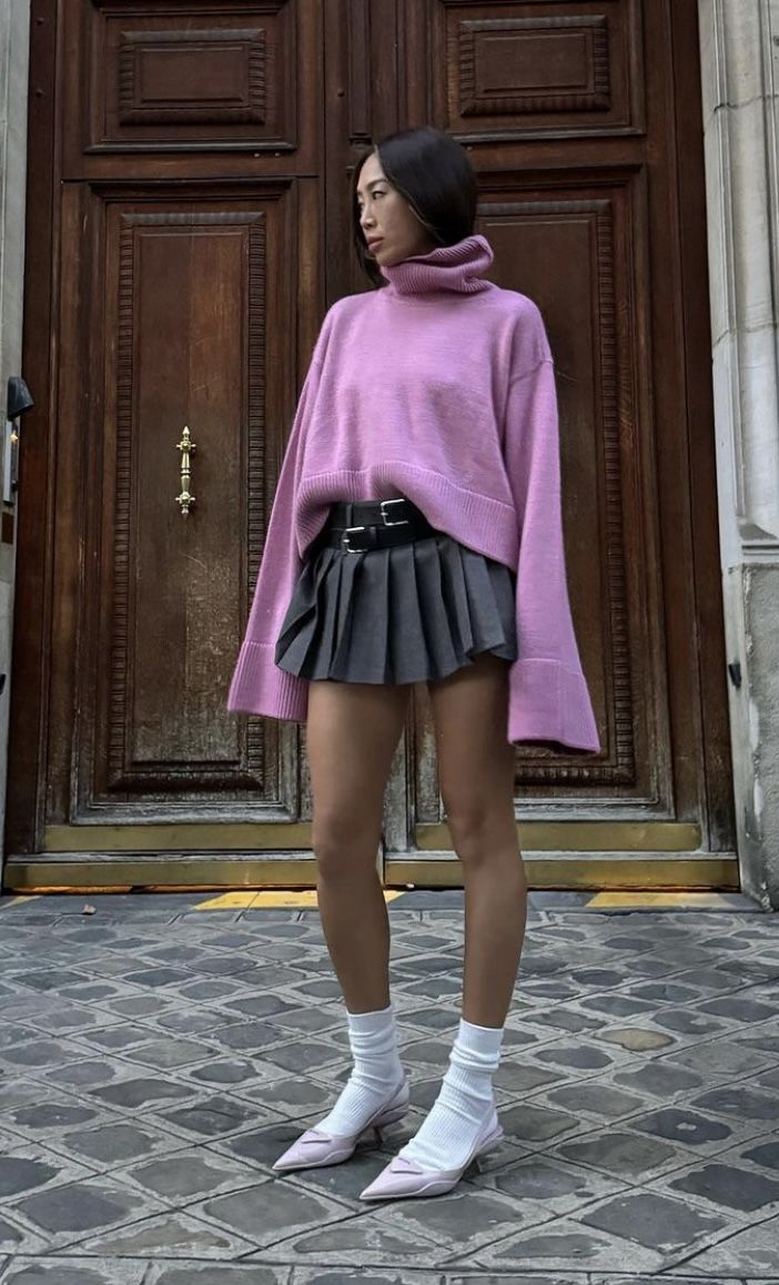 Mulher asi&aacute;tica de cabelo escuro, vestindo um su&eacute;ter rosa de gola alta e saia plissada cinza, posa em frente a uma porta de madeira escura. Ela usa meias brancas e sapatos de salto baixo rosa claro, com o olhar direcionado para a direita. O ch&atilde;o &eacute; de paralelep&iacute;pedos.