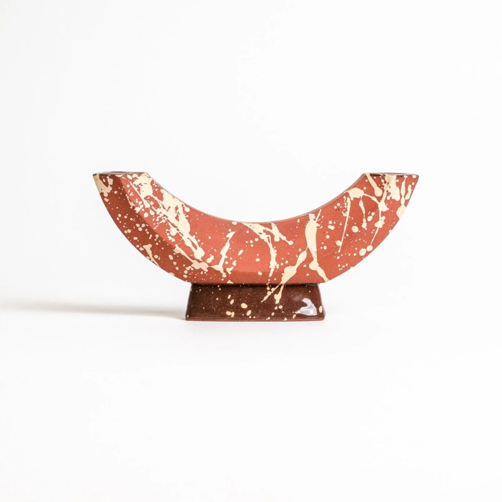 Casti&ccedil;al em formato de meia-lua, terracota com respingos brancos e bege, sobre base retangular marrom escura com pontos brancos, em fundo branco