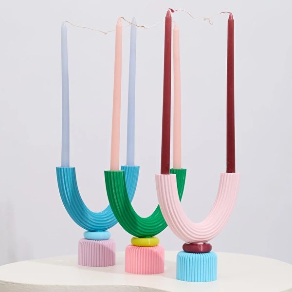 Tr&ecirc;s casti&ccedil;ais coloridos em formato de U invertido, com velas longas. O primeiro &eacute; azul-claro com base rosa e vela cinza. O segundo &eacute; verde com base rosa e vela p&ecirc;ssego. O terceiro &eacute; rosa com base azul e vela vinho.