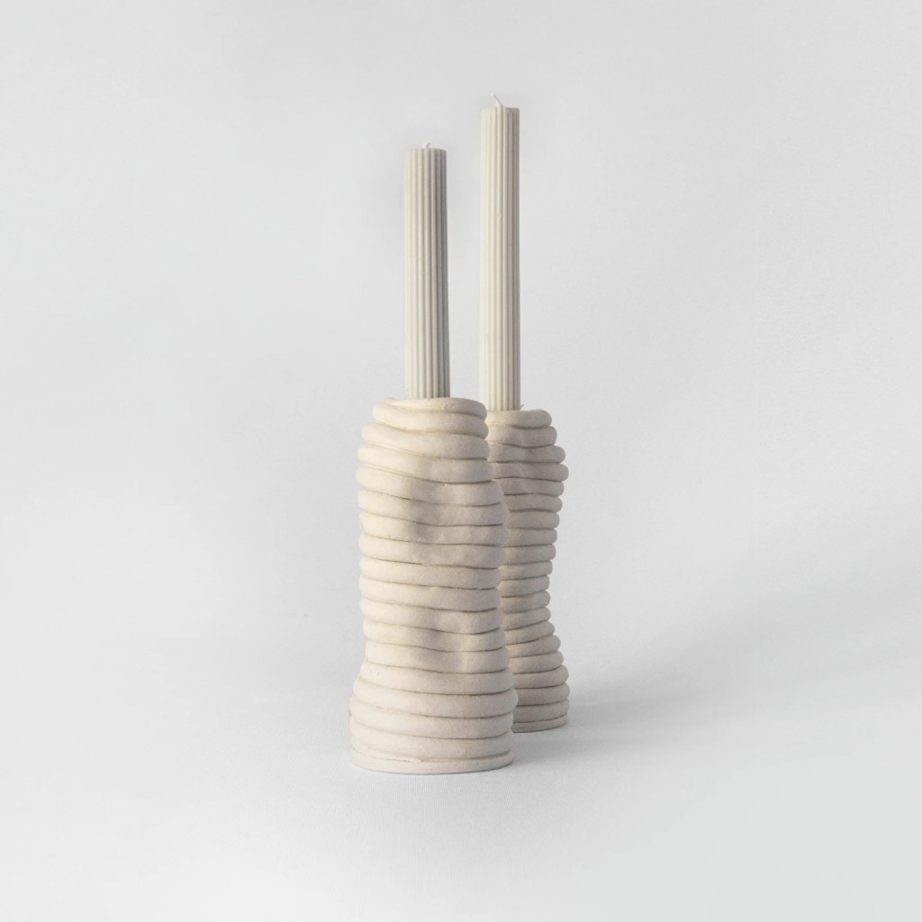 Duas velas cil&iacute;ndricas de cera clara, com textura canelada, inseridas em casti&ccedil;ais de cer&acirc;mica bege com formato org&acirc;nico e an&eacute;is horizontais, sobre fundo branco