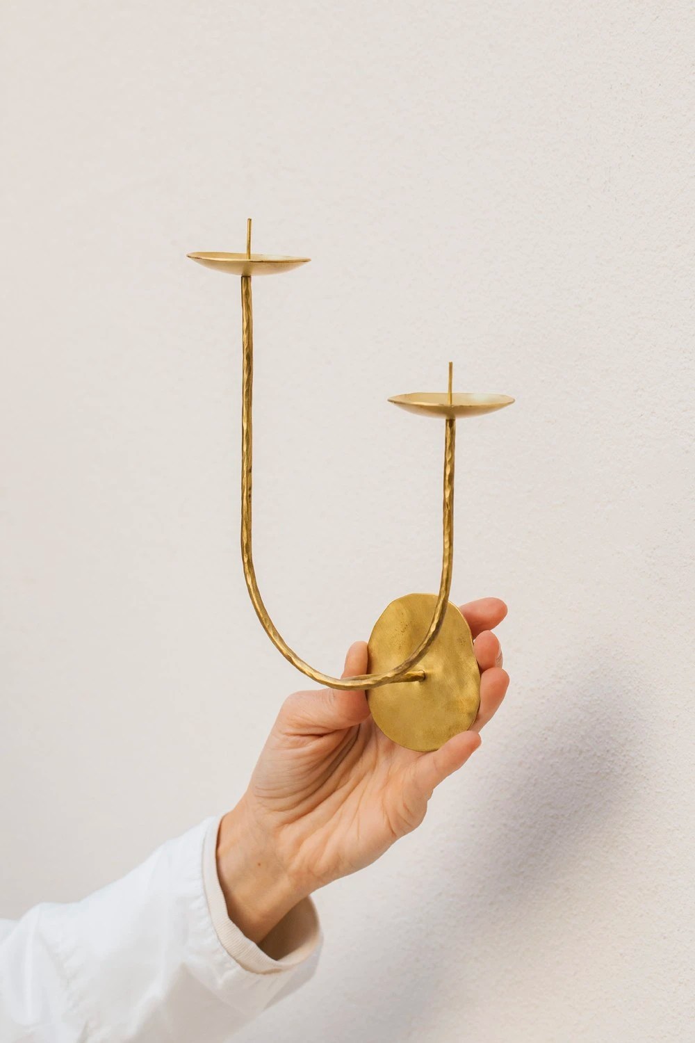 M&atilde;o feminina segura um casti&ccedil;al de parede dourado com dois suportes para velas, em formato de U invertido, contra uma parede branca