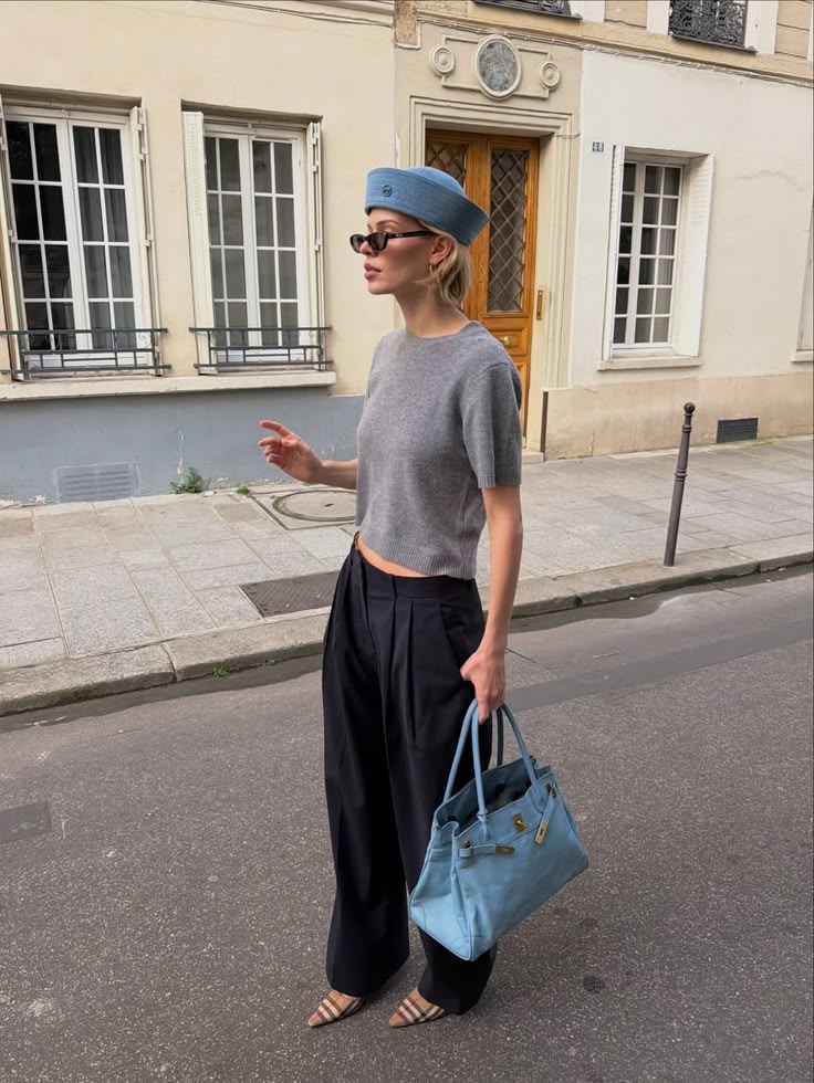 Mulher loira de &oacute;culos escuros, chap&eacute;u azul, blusa cinza e cal&ccedil;a preta larga, segurando uma bolsa azul, caminhando na rua com pr&eacute;dios ao fundo