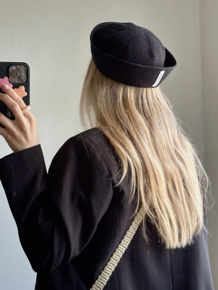 Pessoa de costas com cabelos loiros longos, usando um gorro preto e blazer listrado, segurando um celular com capa decorada e anel no dedo
