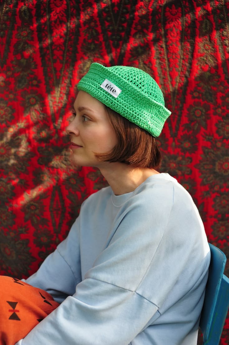 Mulher jovem de cabelo castanho curto, usando um gorro de croch&ecirc; verde-lim&atilde;o com a etiqueta t&ecirc;te e um moletom azul claro, sentada de perfil contra um fundo de tecido vermelho estampado