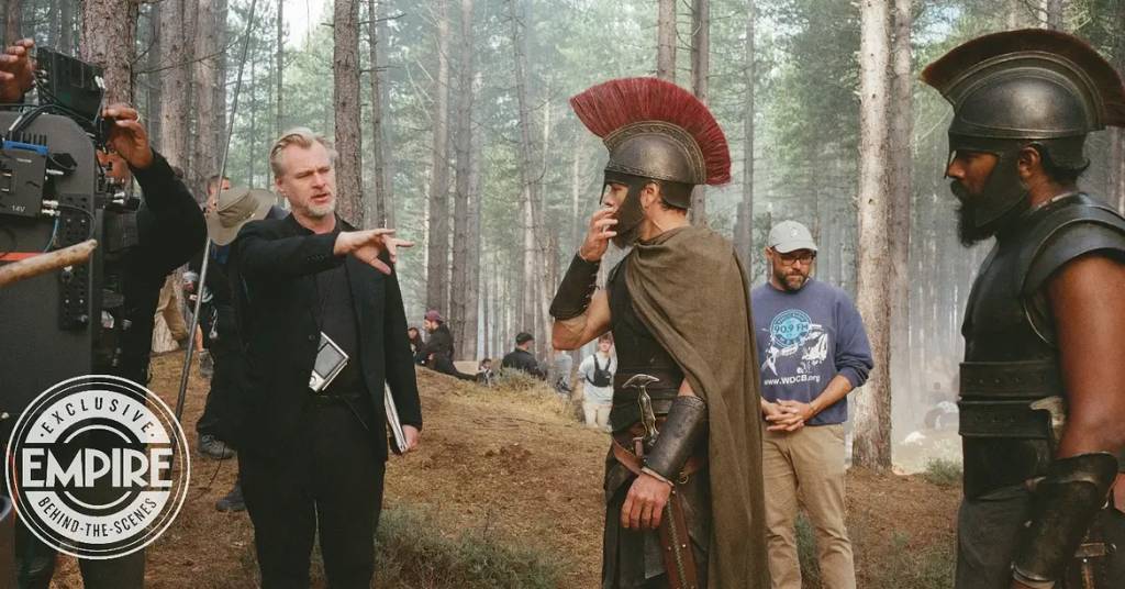 O diretor Christopher Nolan, de terno preto, gesticula enquanto conversa com dois atores vestidos como guerreiros espartanos, um com elmo e capa, e outro com armadura escura, em uma floresta com &aacute;rvores altas e neblina. Um operador de c&acirc;mera e outros membros da equipe est&atilde;o ao redor. O selo Exclusive Empire Behind-the-Scenes aparece no canto inferior esquerdo