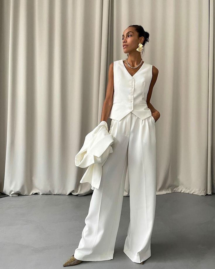 Mulher de pele morena, cabelo preso, vestindo colete e cal&ccedil;a pantalona brancos, com blazer branco dobrado no bra&ccedil;o esquerdo. Ela usa brincos grandes dourados, colar de corrente prateada e sapatos de bico fino dourados, posando em frente a uma cortina bege