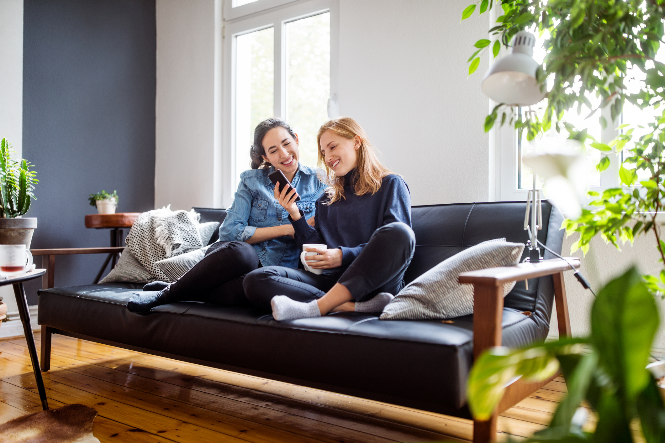 Duas mulheres sorrindo, sentadas em um sofá preto na sala de estar, olhando um celular. Uma veste camisa jeans e a outra blusa azul escura, segurando uma caneca branca. Plantas e uma luminária decoram o ambiente claro