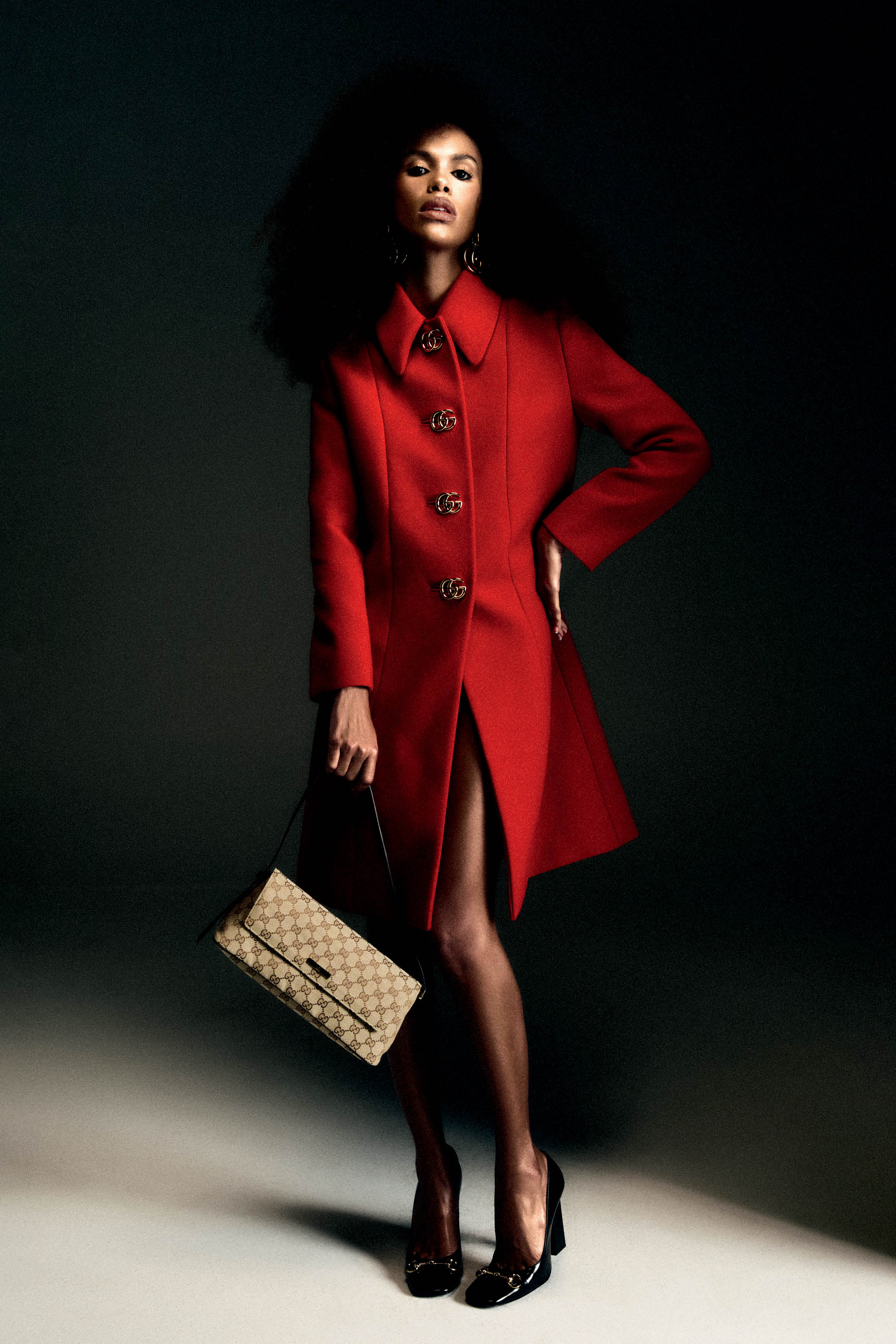 Modelo de pele escura com cabelo afro volumoso, vestindo um casaco vermelho com bot&otilde;es dourados e segurando uma bolsa bege estampada com o monograma GG da Gucci, posando em fundo escuro