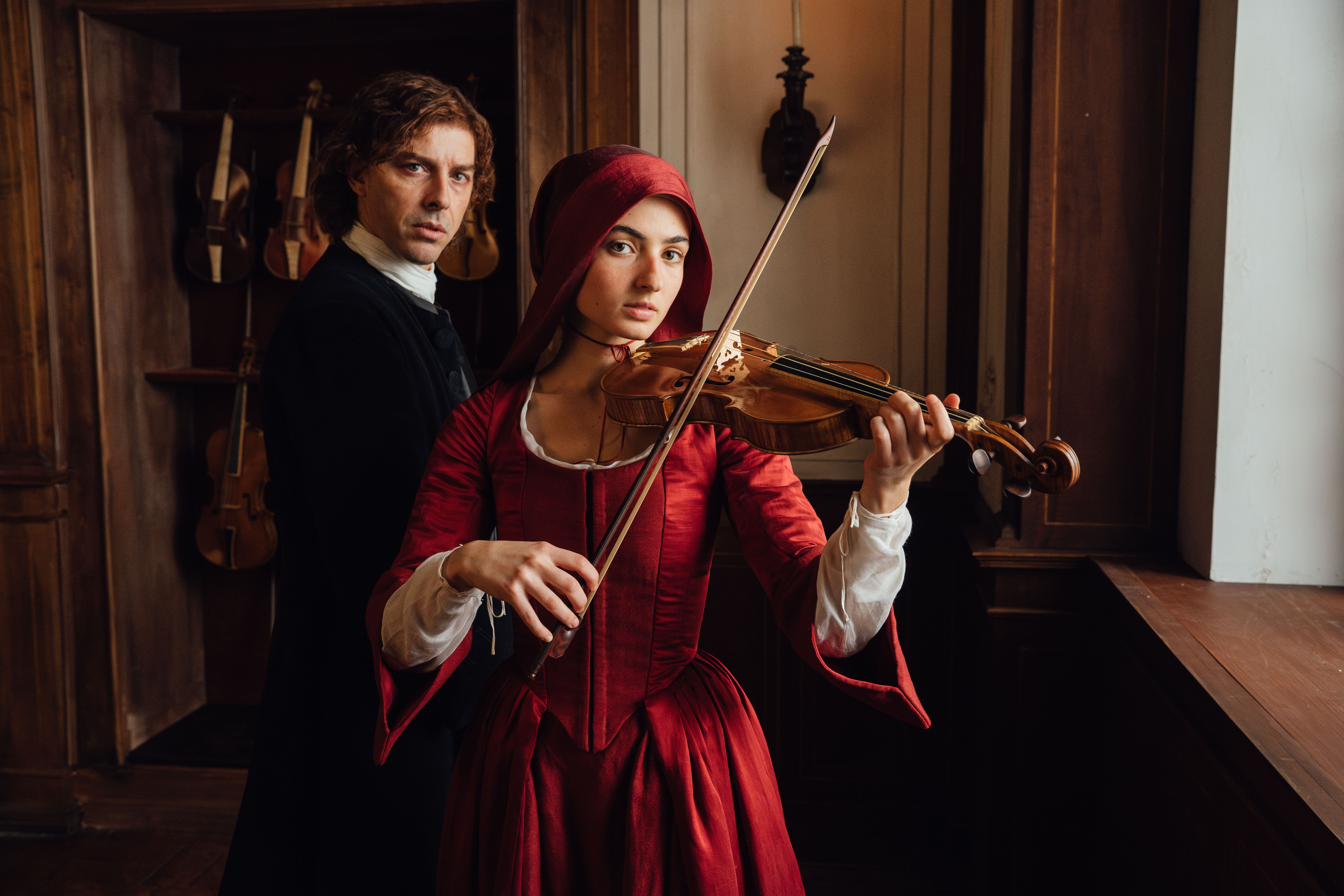 Uma mulher jovem com capuz e vestido vermelho antigo toca violino, olhando fixamente para frente. Atrás dela, um homem de cabelo encaracolado e roupa escura a observa, com violinos pendurados na parede ao fundo