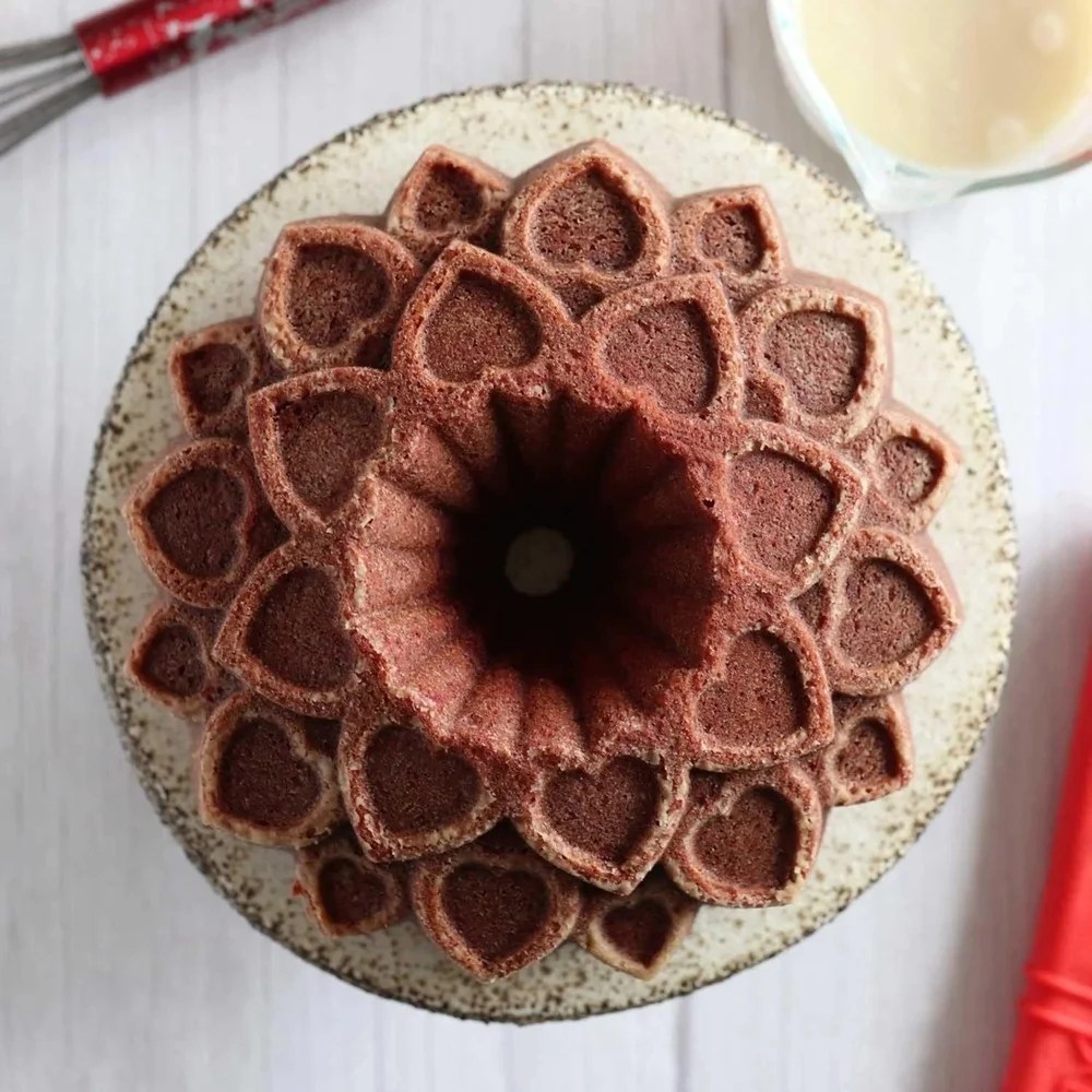 Bolo em formato de flor com cora&ccedil;&otilde;es em relevo, marrom-avermelhado, sobre um prato r&uacute;stico. Ao fundo, um fouet e uma tigela com l&iacute;quido claro.