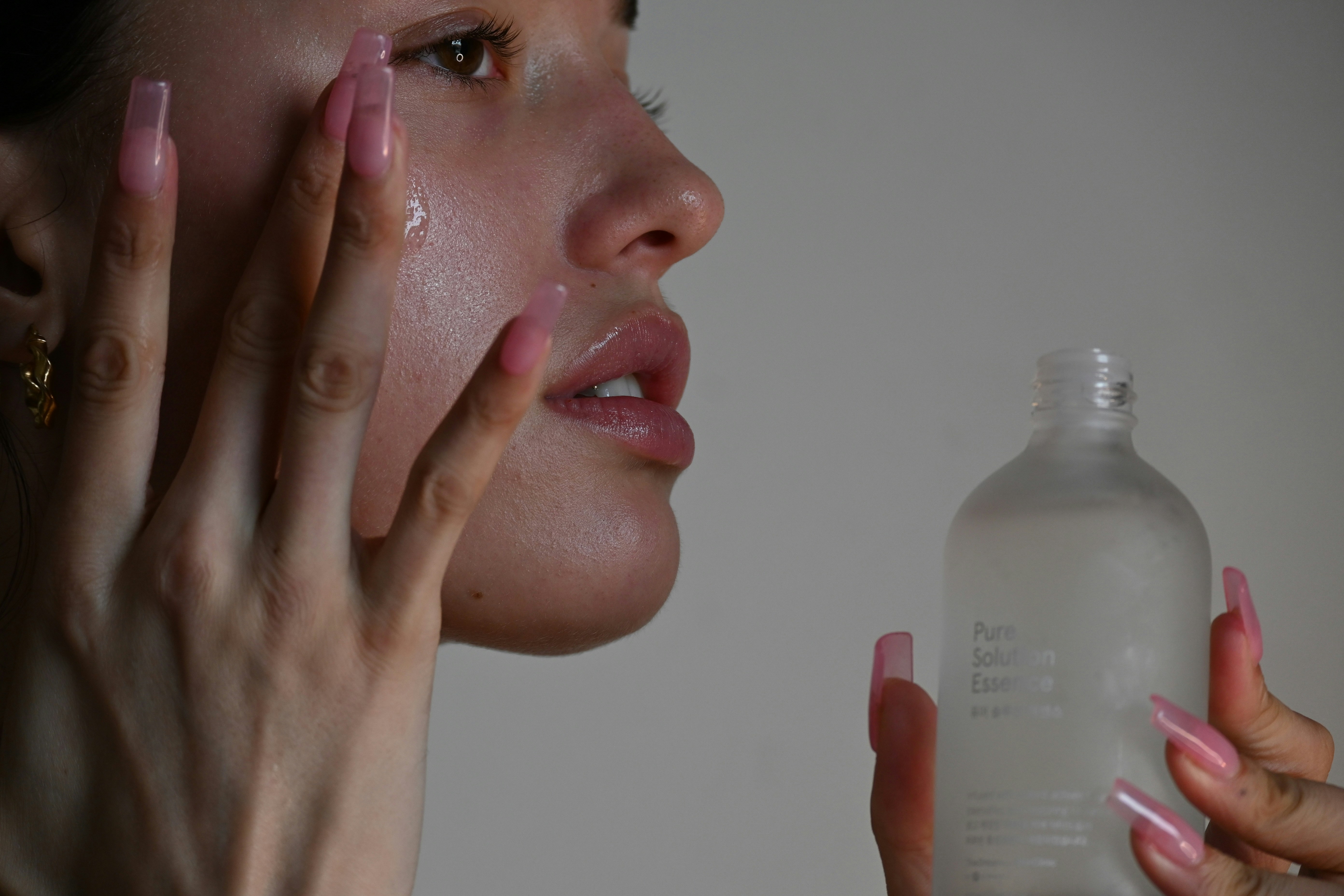 Mulher com unhas longas e rosadas aplicando um produto l&iacute;quido transparente no rosto, que escorre pela bochecha. Ela segura um frasco branco fosco com a m&atilde;o direita, com a inscri&ccedil;&atilde;o Pure Solution Essence vis&iacute;vel. O fundo &eacute; cinza claro