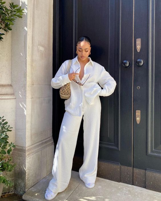 Mulher de pele morena, cabelo preso, vestindo conjunto branco texturizado de camisa e cal&ccedil;a larga, segurando &oacute;culos escuros. Ela est&aacute; em frente a uma porta preta, com uma bolsa bege de estampa marrom no ombro e t&ecirc;nis brancos. &Agrave; esquerda, um pilar claro e folhagem verde.