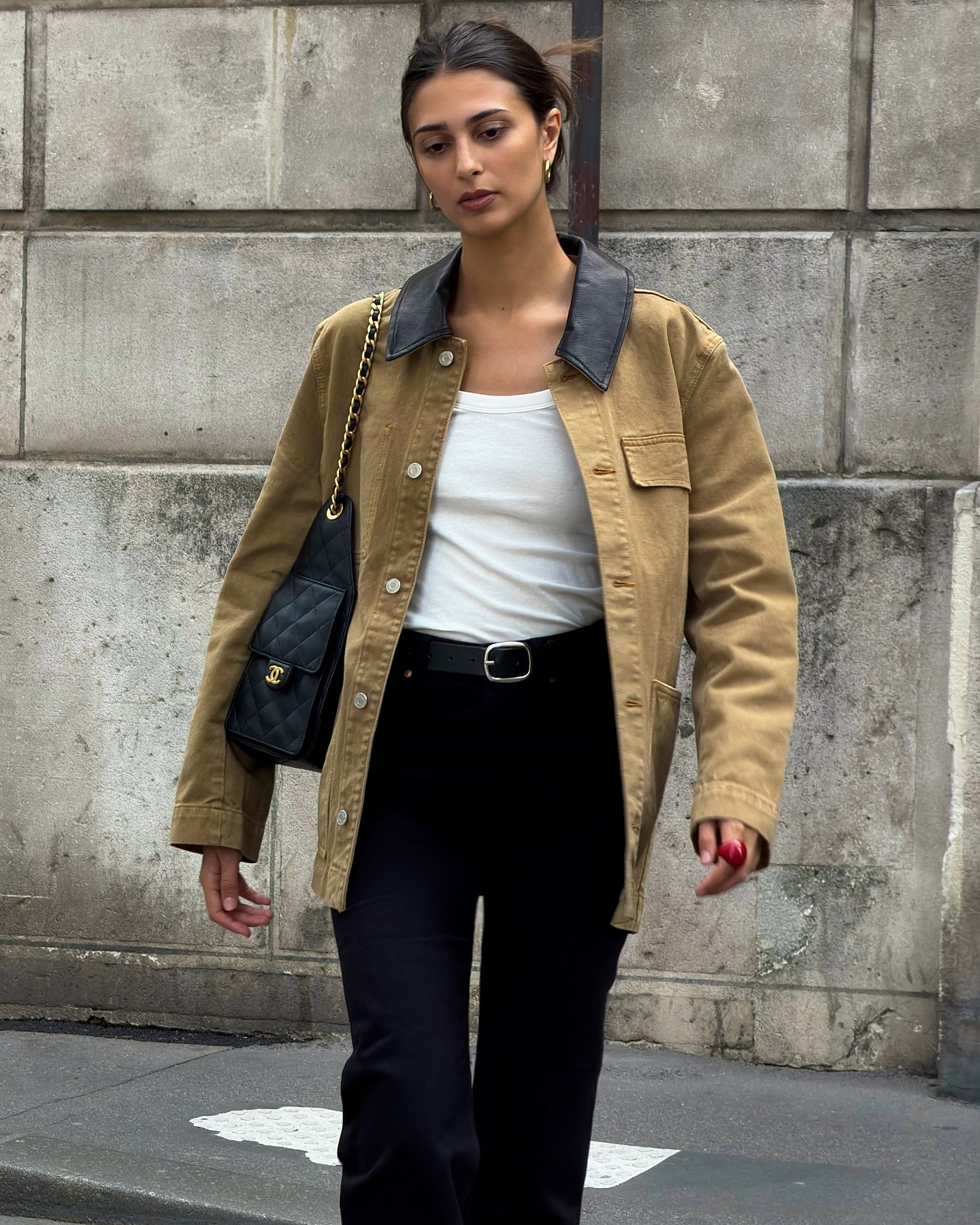 Mulher jovem com cabelo castanho preso, vestindo jaqueta c&aacute;qui com gola preta, regata branca, cal&ccedil;a preta e cinto preto, caminhando em frente a uma parede de blocos de concreto. Ela carrega uma bolsa preta acolchoada com al&ccedil;a de corrente dourada no ombro esquerdo e usa um anel vermelho no dedo anelar direito