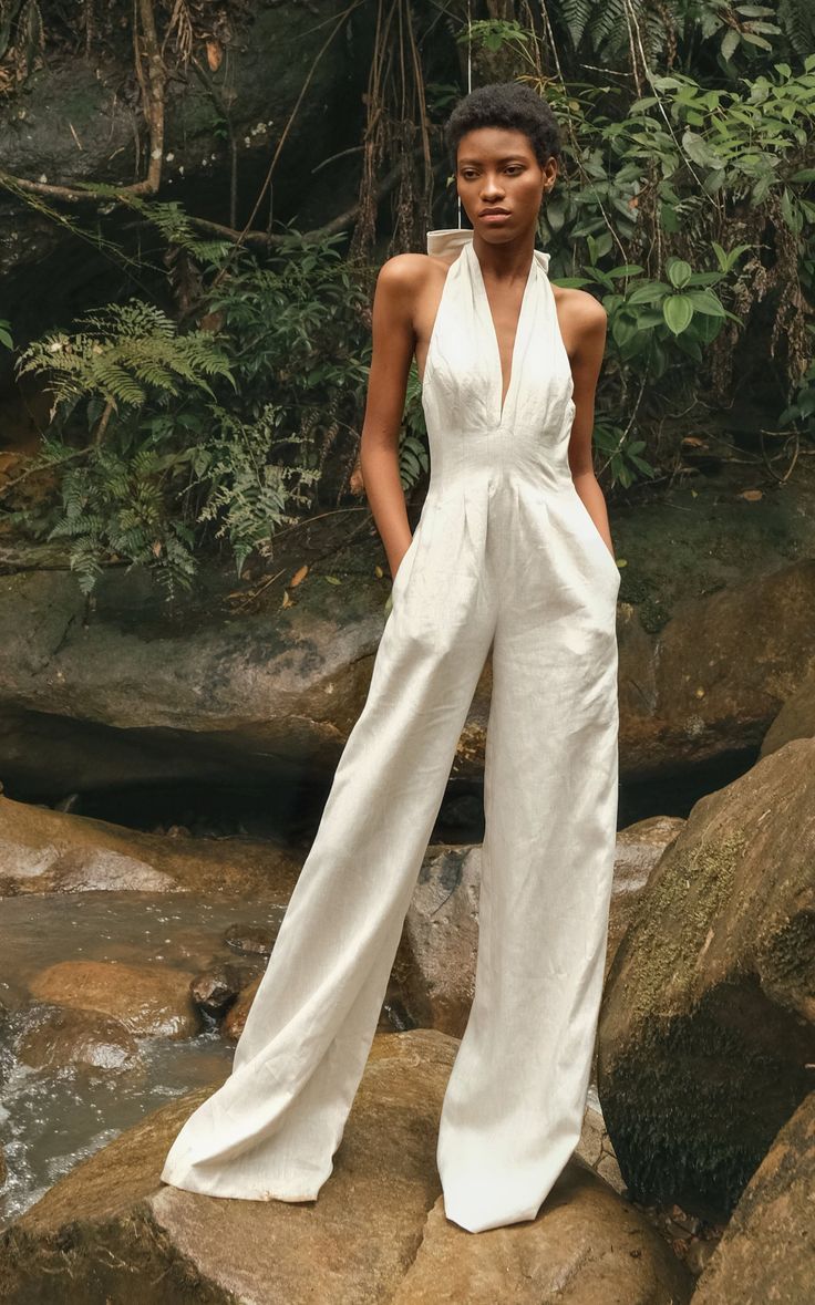 Mulher negra de cabelo curto e crespo, vestindo um macac&atilde;o branco de linho com decote V e al&ccedil;as largas, posa com as m&atilde;os nos bolsos em um riacho com pedras e vegeta&ccedil;&atilde;o exuberante