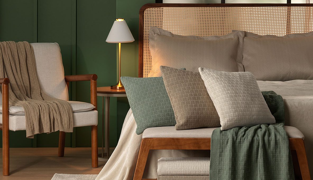 Quarto com parede verde-escura, cama com cabeceira de palha e roupa de cama bege. Na frente da cama, um banco de madeira com almofadas e manta verde. À esquerda, poltrona clara com manta marrom e mesa de cabeceira com abajur aceso. No fundo, janela com vidro fosco e grades pretas