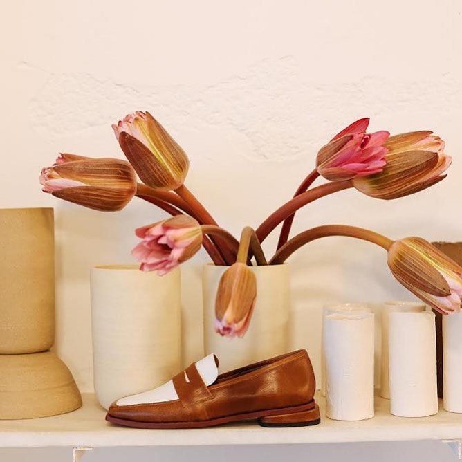 Um mocassim marrom e branco sobre uma prateleira branca, com um vaso de cer&acirc;mica contendo flores de l&oacute;tus rosadas e fechadas ao fundo, e outros objetos decorativos cil&iacute;ndricos em tons neutros
