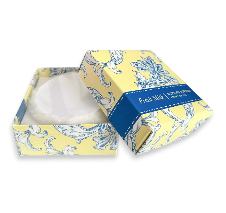 Caixa amarela e azul com estampa floral barroca, aberta, revelando um aplicador de p&oacute; branco. A tampa tem uma faixa azul com Fresh Milk Dusting Powder e NET WT. 4oz 113g em branco