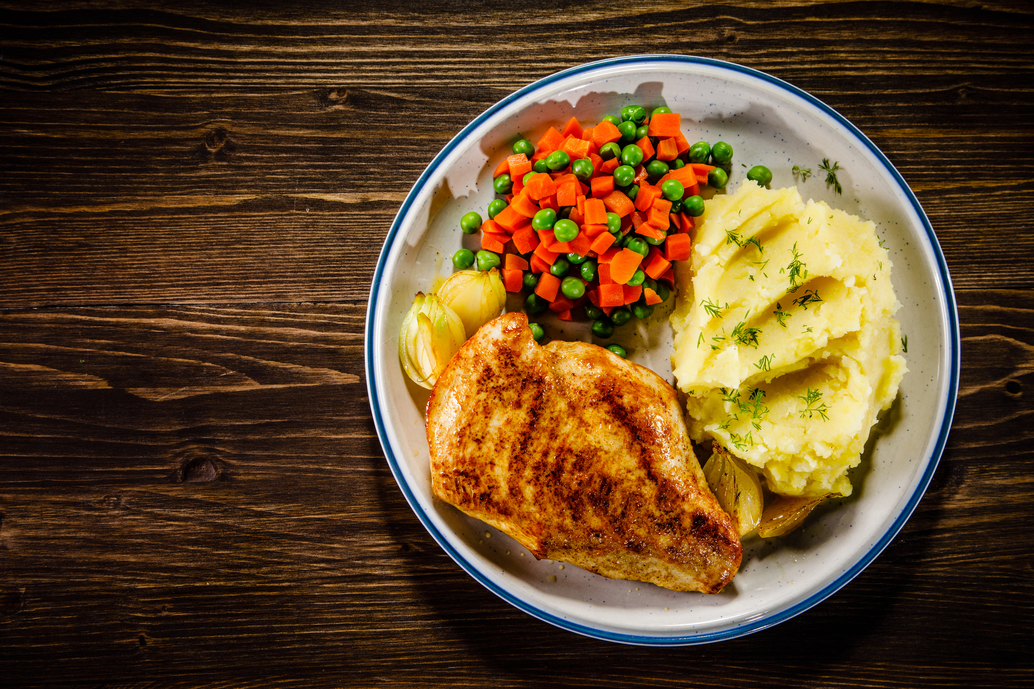 Prato com peito de frango grelhado, pur&ecirc; de batatas com ervas, ervilhas e cenouras picadas, e cebolas assadas, sobre uma mesa de madeira escura