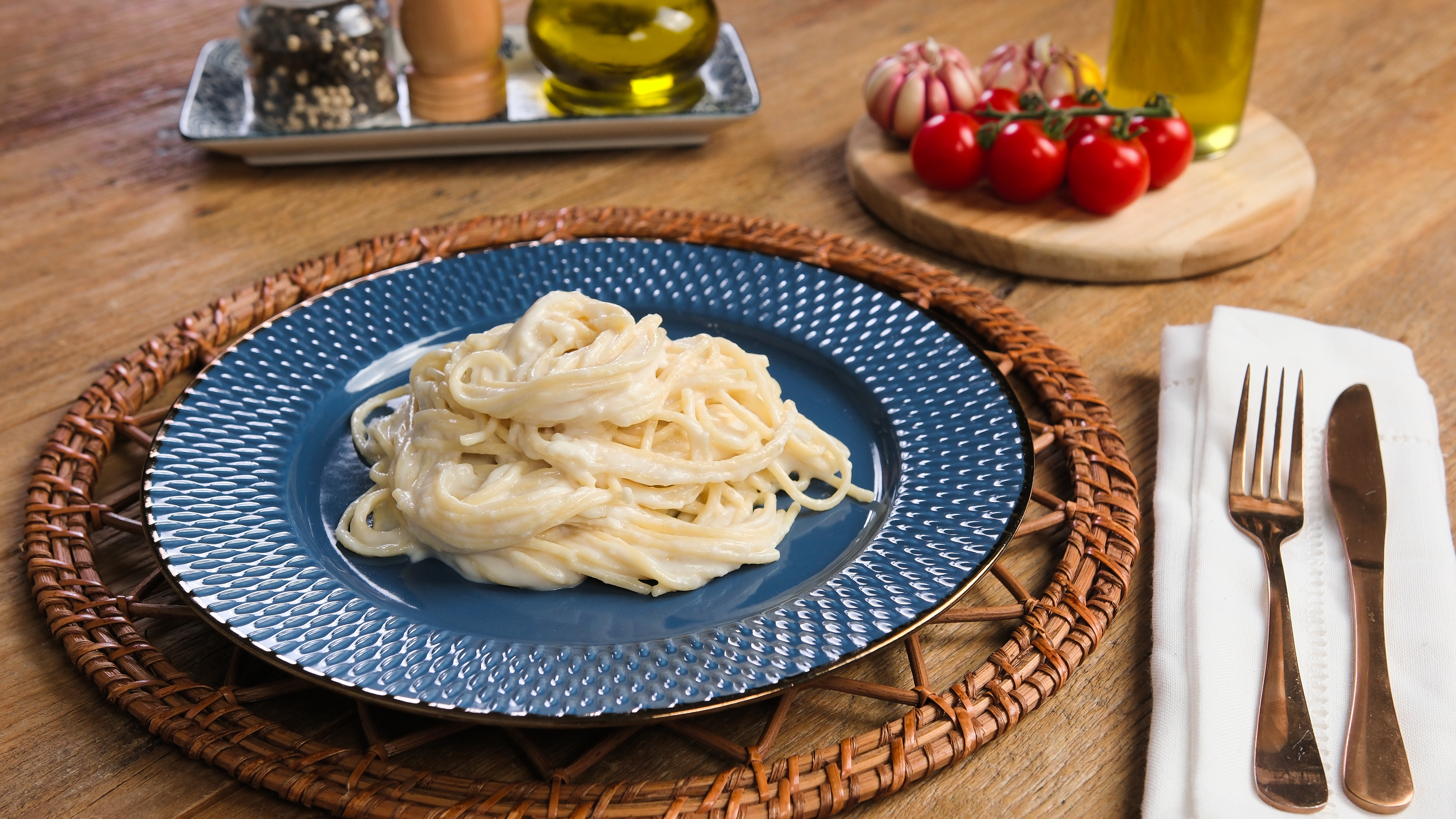 Prato azul com espaguete cremoso sobre um sousplat de vime, acompanhado de talheres de cobre em guardanapo branco, alho, tomates e azeite em uma mesa de madeira