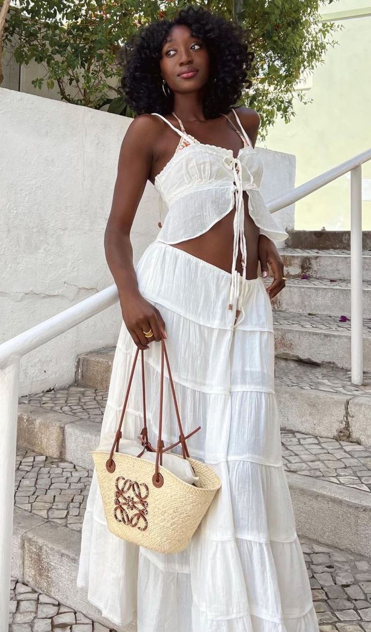 Mulher negra com cabelo cacheado, vestindo um conjunto branco de top cropped e saia longa com babados, segurando uma bolsa de palha com al&ccedil;as de couro marrom, em uma escadaria de pedra com corrim&atilde;o branco e vegeta&ccedil;&atilde;o ao fundo