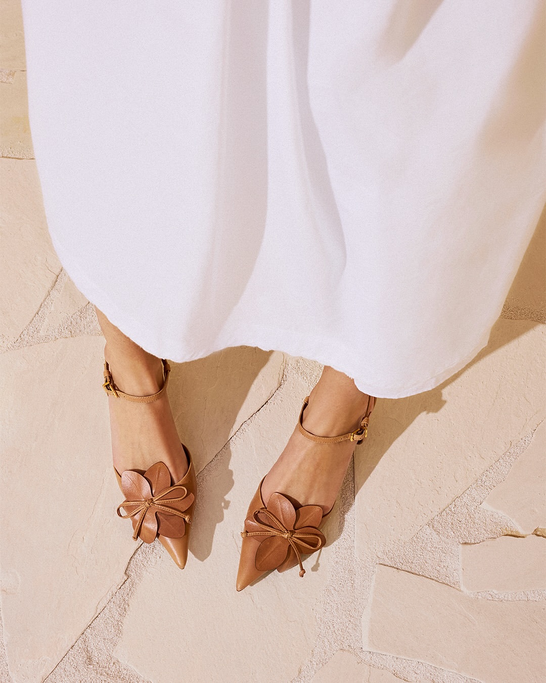 P&eacute;s de uma pessoa com vestido branco, usando sapatilhas de bico fino cor caramelo, com tiras no tornozelo e detalhes de flor e la&ccedil;o na frente, sobre um piso de pedras claras