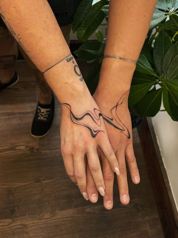 Duas m&atilde;os, uma masculina e outra feminina, com tatuagens combinando de linhas onduladas pretas que se encontram no centro. Ambas usam pulseiras finas de corrente prateada, e a m&atilde;o feminina tem unhas pintadas em tom nude. Ao fundo, um piso de madeira e uma planta verde