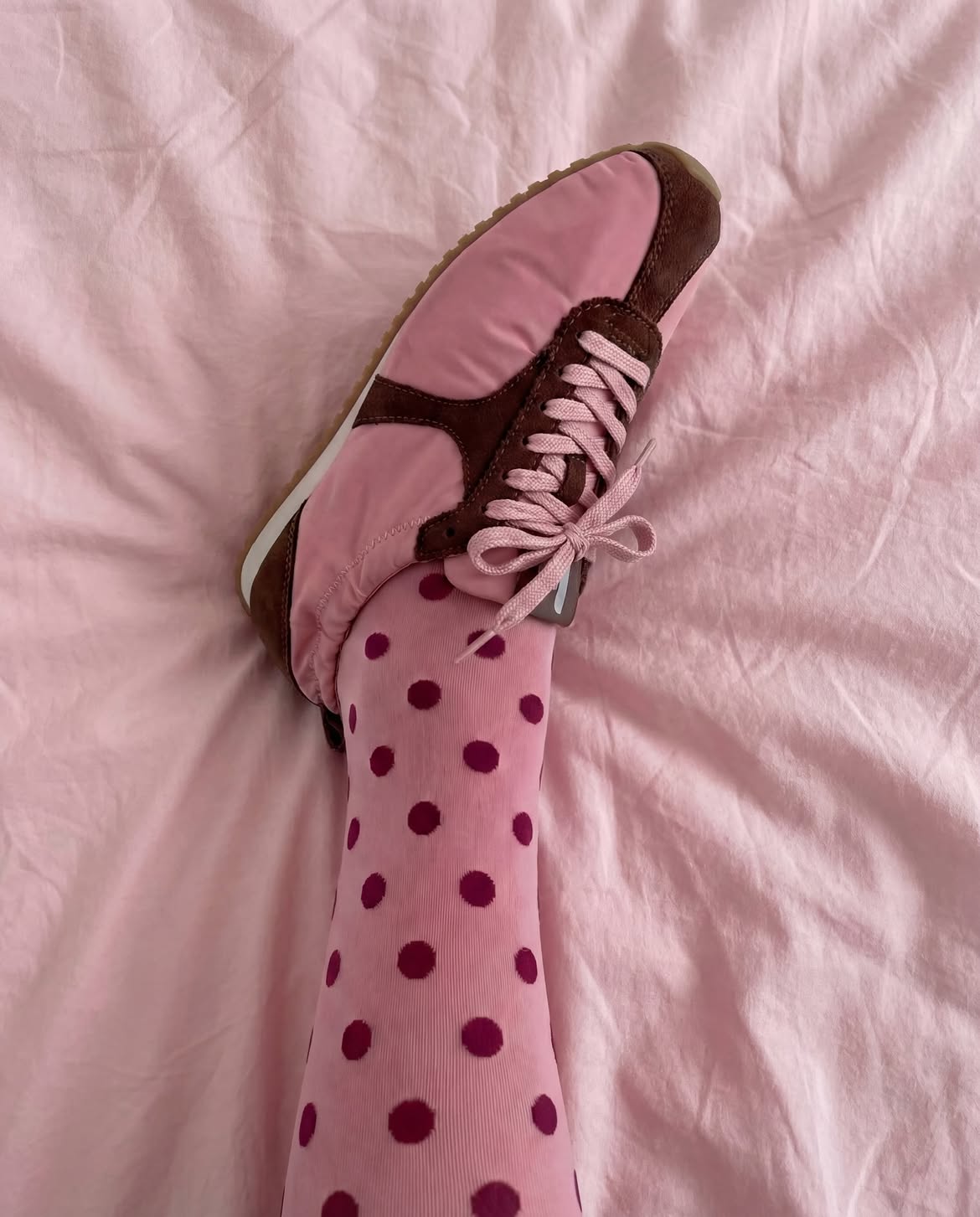 Perna com meia-cal&ccedil;a rosa de bolinhas vinho e t&ecirc;nis rosa com detalhes marrons, sobre um len&ccedil;ol rosa claro amassado
