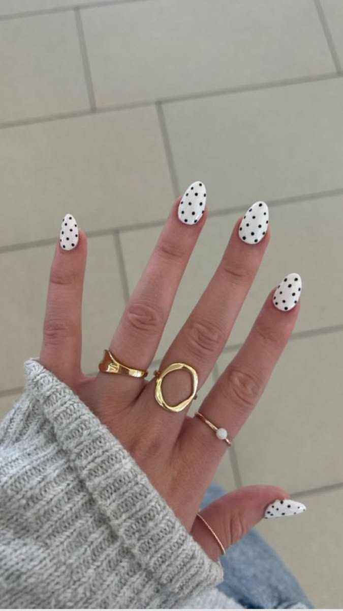 M&atilde;o feminina com unhas ovais brancas e bolinhas pretas, usando an&eacute;is dourados e uma blusa de tric&ocirc; cinza