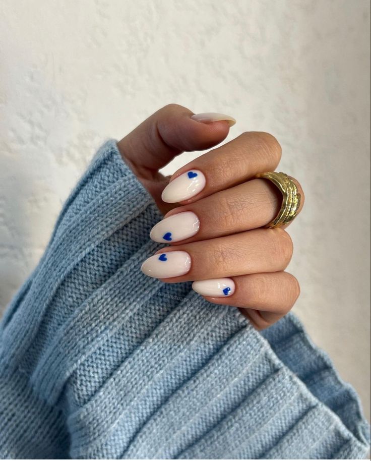 M&atilde;o feminina com unhas ovais brancas, cada uma decorada com um pequeno cora&ccedil;&atilde;o azul. Ela usa um anel dourado no dedo anelar e uma blusa de tric&ocirc; azul claro