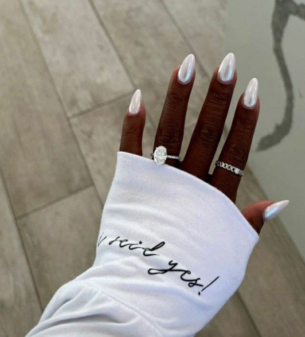 M&atilde;o de pele escura com unhas ovais peroladas, usando anel de noivado com diamante oval e outro anel prateado, sobre manga branca com a frase I said yes! em preto