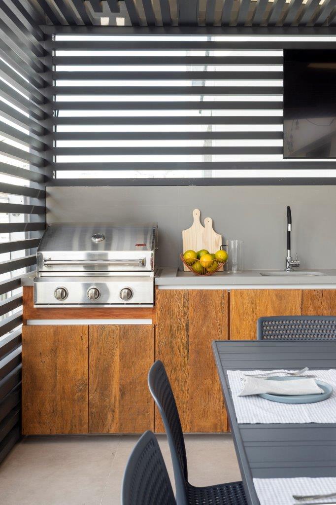 &Aacute;rea gourmet moderna com churrasqueira de inox embutida, bancada cinza clara, arm&aacute;rios de madeira r&uacute;stica e pia com torneira preta. Uma fruteira com lim&otilde;es e t&aacute;buas de corte de madeira decoram a bancada. &Agrave; direita, uma mesa cinza com cadeiras escuras e um lugar posto com prato azul e guardanapo branco. O teto tem ripas de madeira escura e ilumina&ccedil;&atilde;o embutida