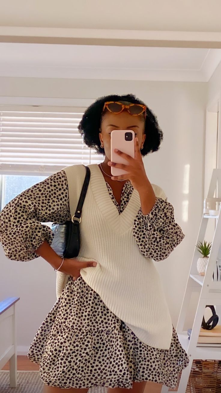 Mulher de pele escura e cabelo crespo, usando &oacute;culos de sol na cabe&ccedil;a, tira selfie no espelho. Veste um vestido estampado de bolinhas pretas e brancas com mangas bufantes, sobreposto por um colete de tric&ocirc; creme. Ela segura uma bolsa preta de al&ccedil;a curta no ombro e um celular rosa claro na frente do rosto. Ao fundo, uma janela com persianas e uma estante branca com plantas e livros