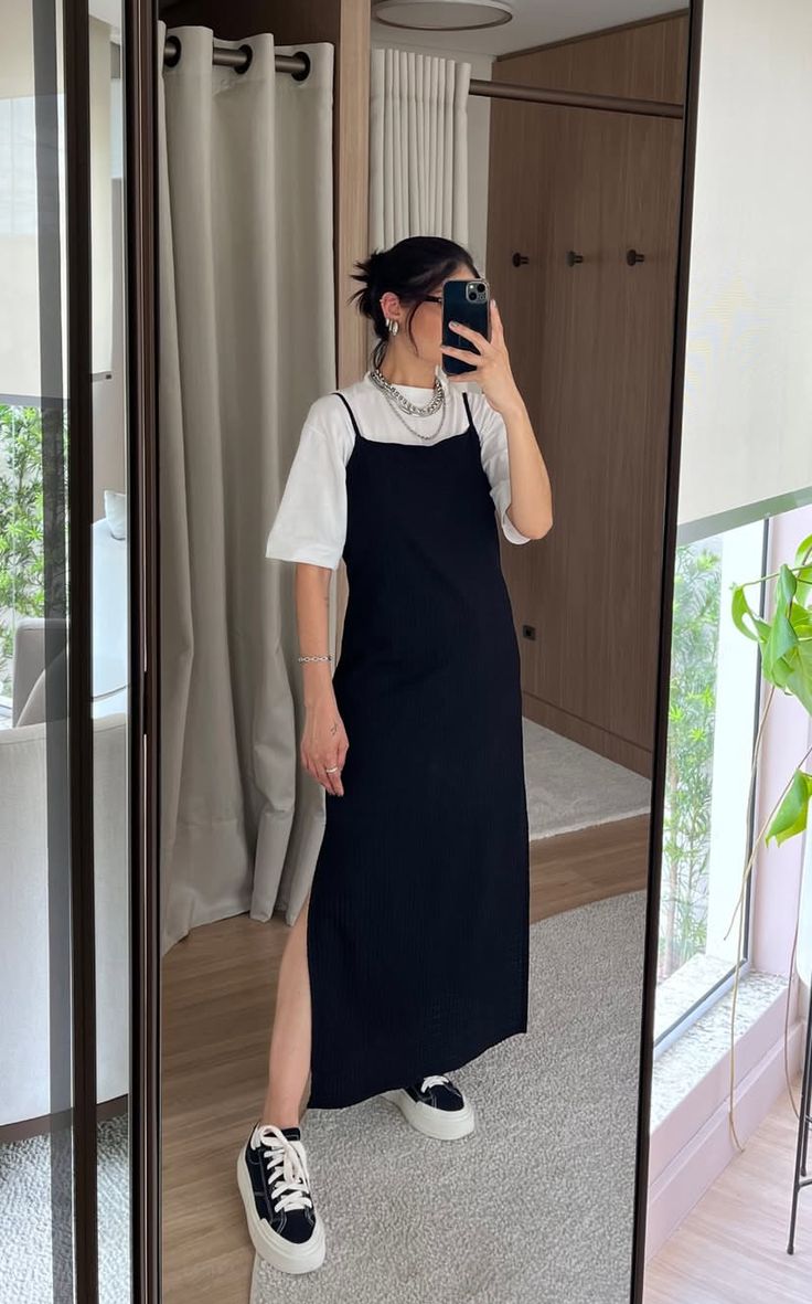 Mulher de cabelo preso, &oacute;culos escuros e camiseta branca sob um vestido preto de al&ccedil;as finas, com fenda lateral, tira uma selfie no espelho. Ela usa t&ecirc;nis pretos com solado branco e acess&oacute;rios prateados, incluindo um colar de correntes e brincos de argola. O ambiente reflete cortinas claras, um arm&aacute;rio de madeira e um vaso de planta verde