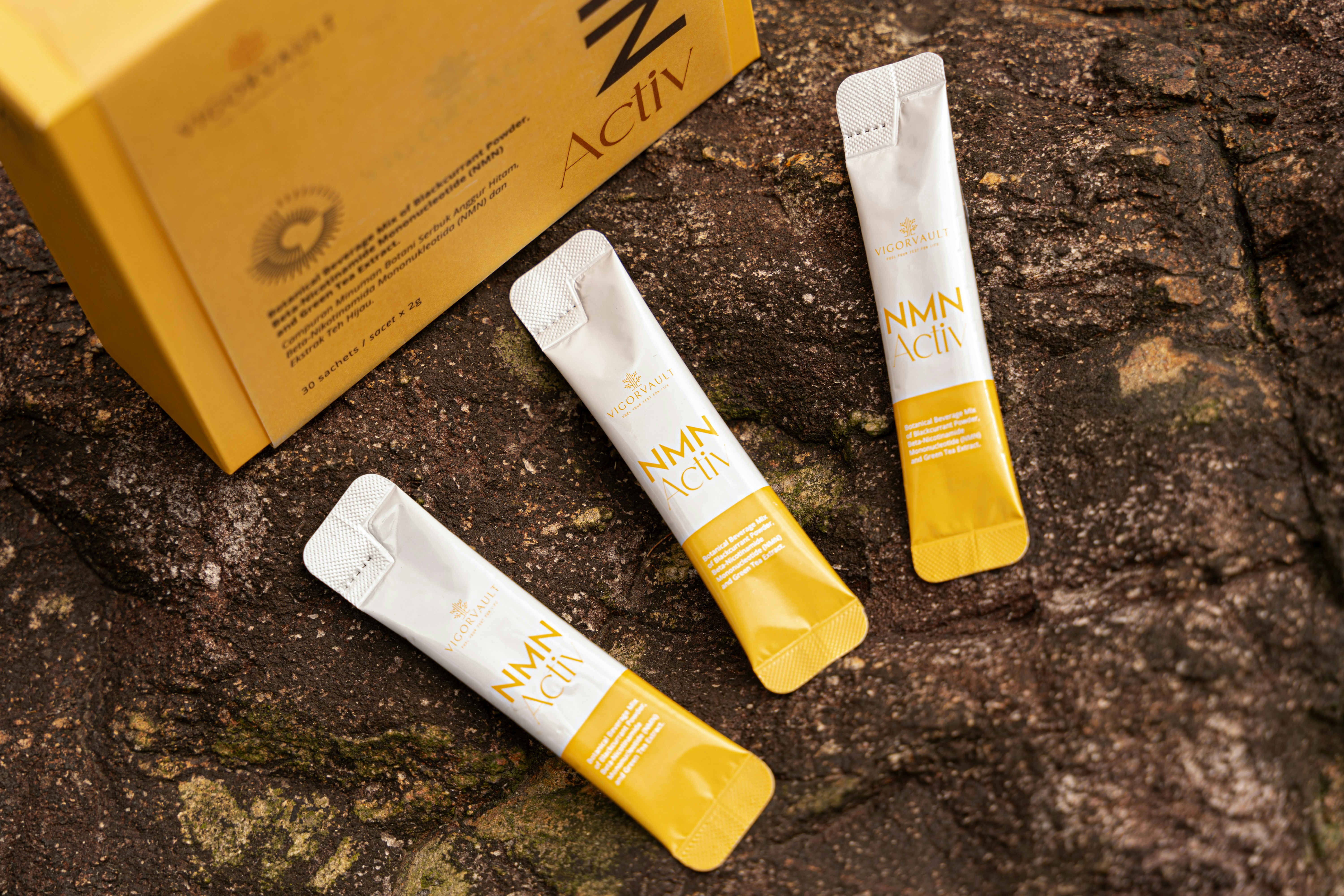 Tr&ecirc;s sach&ecirc;s brancos e amarelos de NMN Activ, da Vigorvault, sobre uma superf&iacute;cie rochosa escura. Uma caixa amarela do produto aparece parcialmente no canto superior esquerdo, com texto em ingl&ecirc;s e indon&eacute;sio sobre 30 sach&ecirc;s de 2g