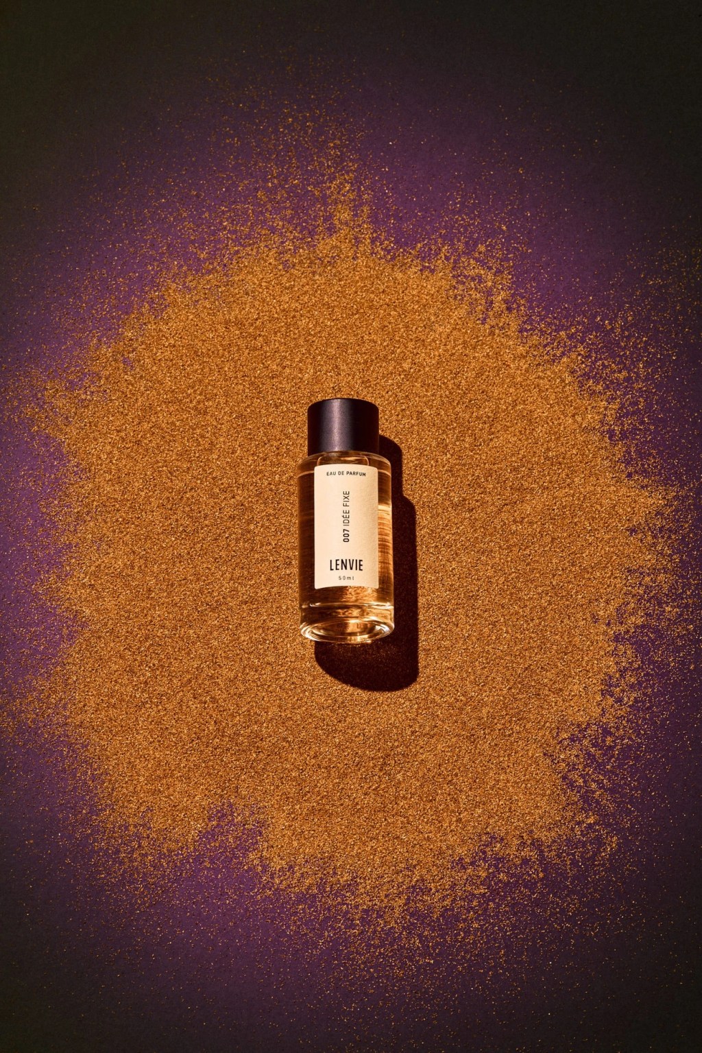 Frasco de perfume Lenvie 007 Id&eacute;e Fixe de 50ml, com l&iacute;quido &acirc;mbar e tampa preta, sobre uma superf&iacute;cie roxa escura coberta por glitter dourado em formato de nuvem