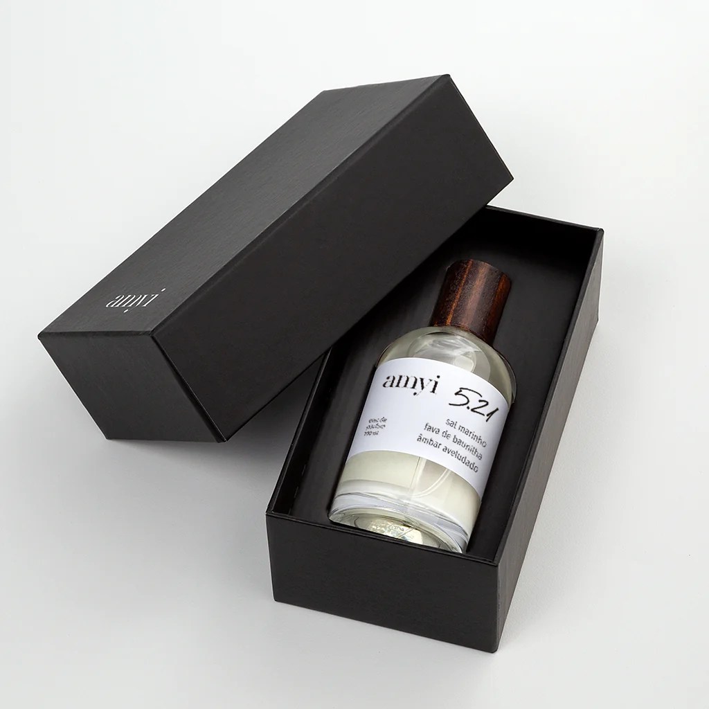 Frasco de perfume Amyi 5.21 com tampa de madeira escura dentro de uma caixa preta aberta, sobre fundo branco