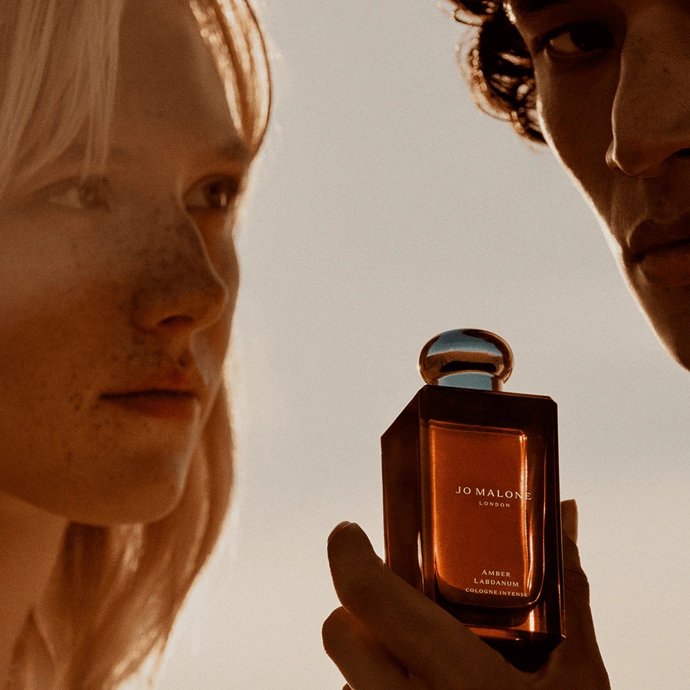 Um homem e uma mulher, com a pele iluminada por um p&ocirc;r do sol dourado, observam um frasco de perfume Jo Malone London Amber & Labdanum Cologne Intense, segurado pela m&atilde;o do homem. O frasco retangular tem l&iacute;quido &acirc;mbar e tampa esf&eacute;rica prateada.
