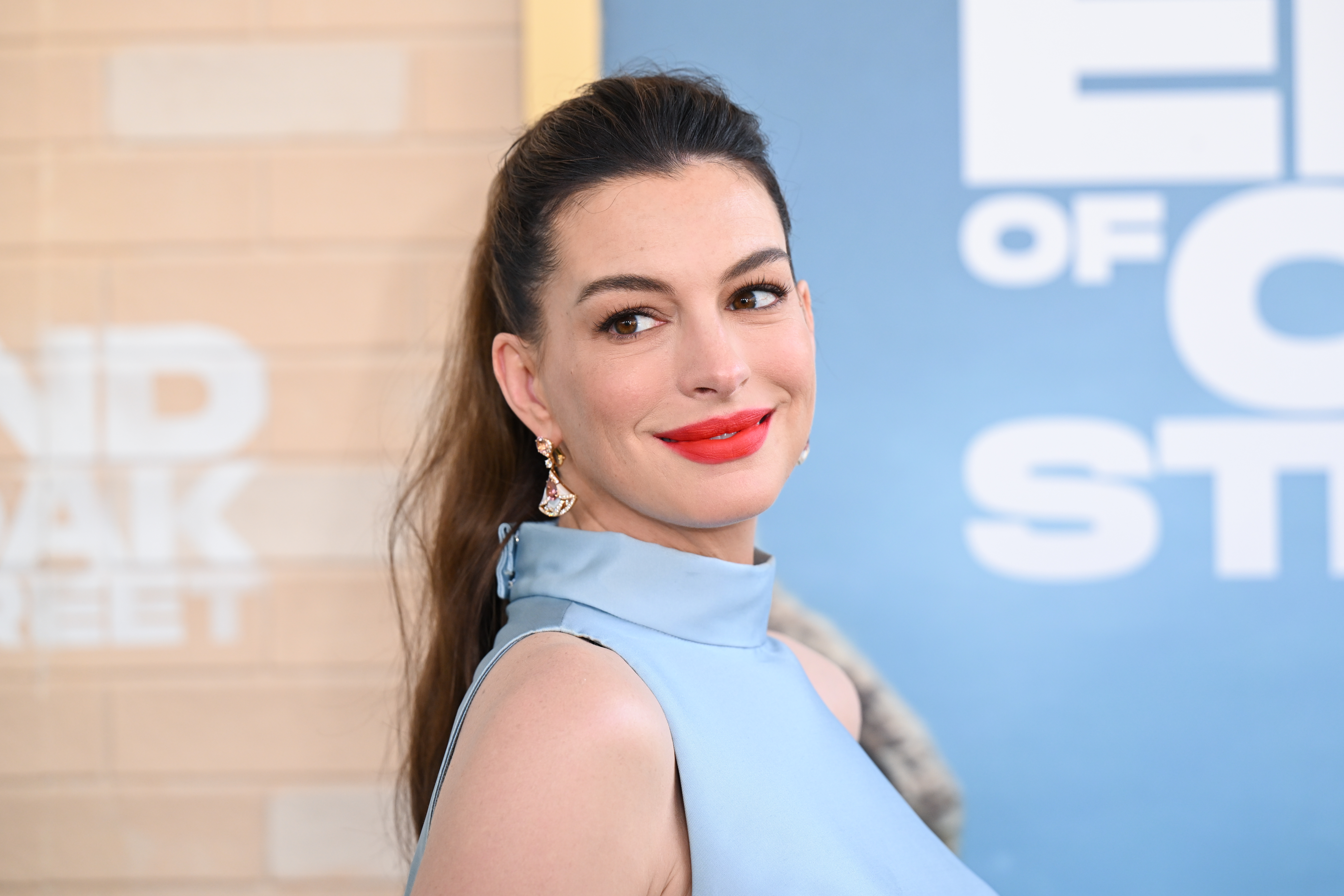 Anne Hathaway sorrindo, com cabelo castanho preso em rabo de cavalo, maquiagem leve e batom vermelho. Ela usa um vestido azul-claro de gola alta e brincos dourados com detalhes coloridos. Ao fundo, uma parede de tijolos claros e um banner azul com letras brancas desfocadas