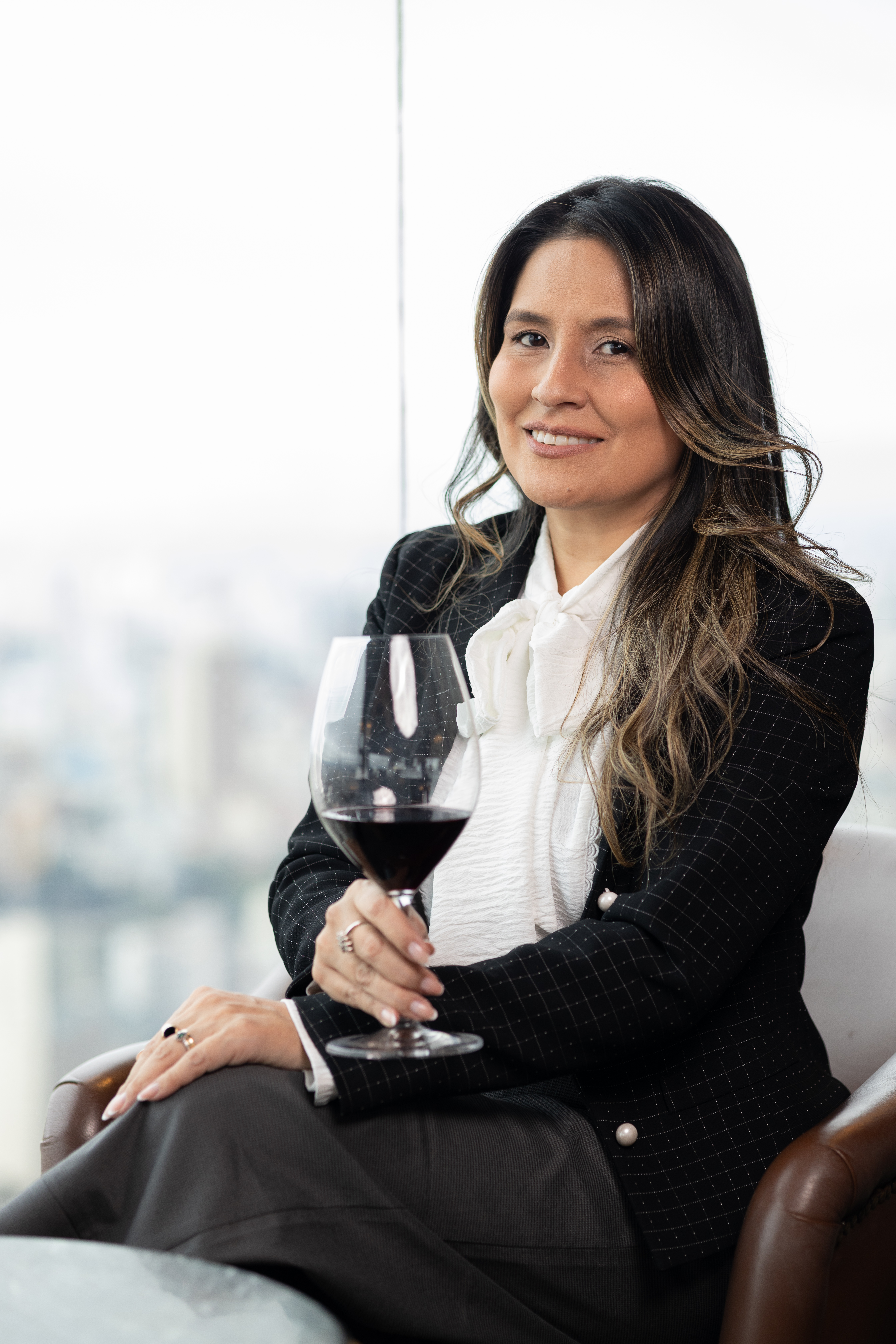 Mulher sorridente, de cabelos castanhos longos, sentada em poltrona de couro, segurando uma ta&ccedil;a de vinho tinto. Ela veste blazer preto xadrez, blusa branca com la&ccedil;o e cal&ccedil;a escura, com vista para uma cidade ao fundo