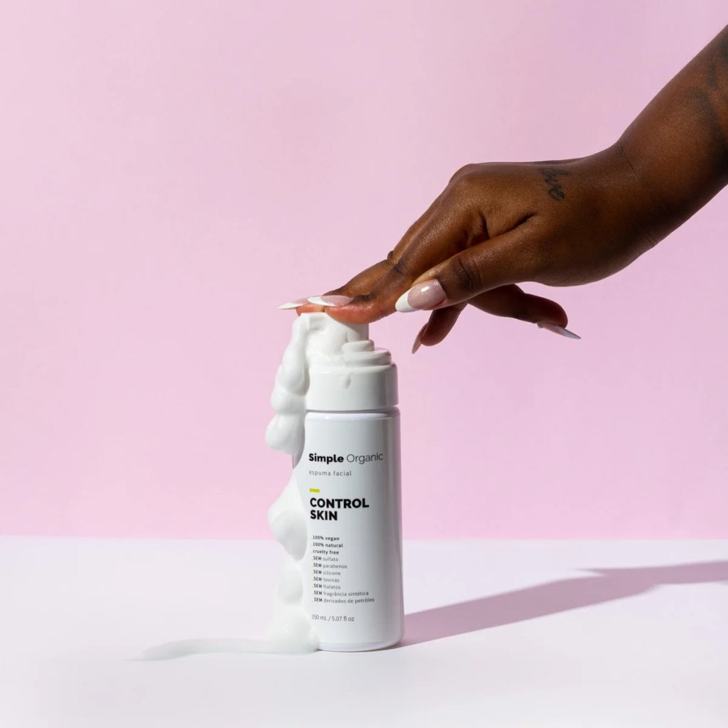 M&atilde;o de pele escura com unhas francesinhas pressiona o pump de um frasco branco de espuma facial Simple Organic Control Skin, que libera espuma branca escorrendo pelo frasco e base, sobre fundo rosa
