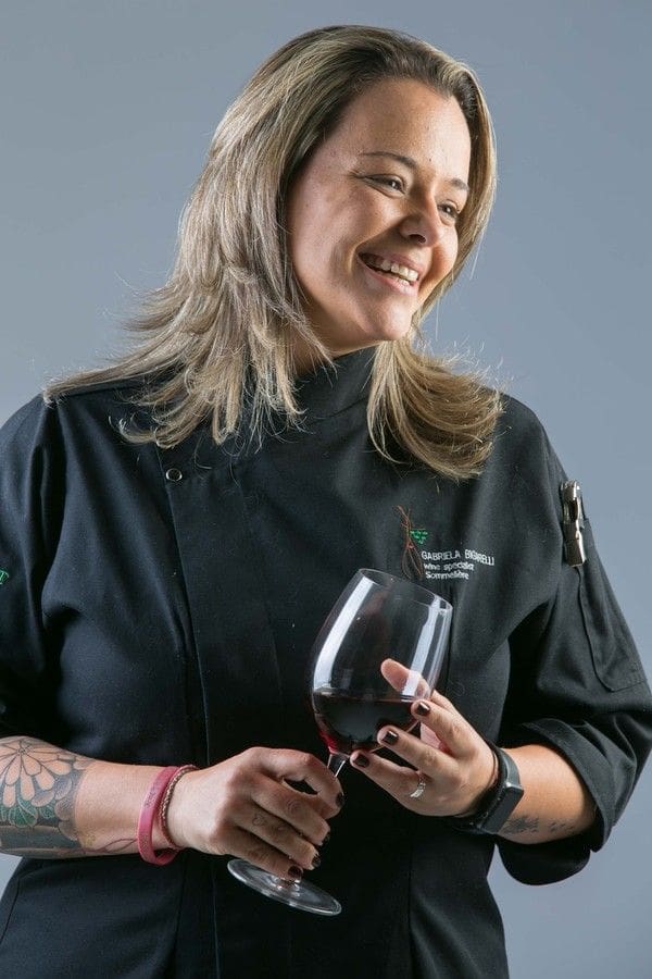 Mulher de cabelos loiros e pele morena, sorrindo, vestindo d&oacute;lm&atilde; preto com logo Gabriela Bigarelli Wine Specialist Sommeli&egrave;re e segurando uma ta&ccedil;a de vinho tinto. Ela tem tatuagem floral no bra&ccedil;o esquerdo e usa pulseiras rosas e rel&oacute;gio inteligente preto
