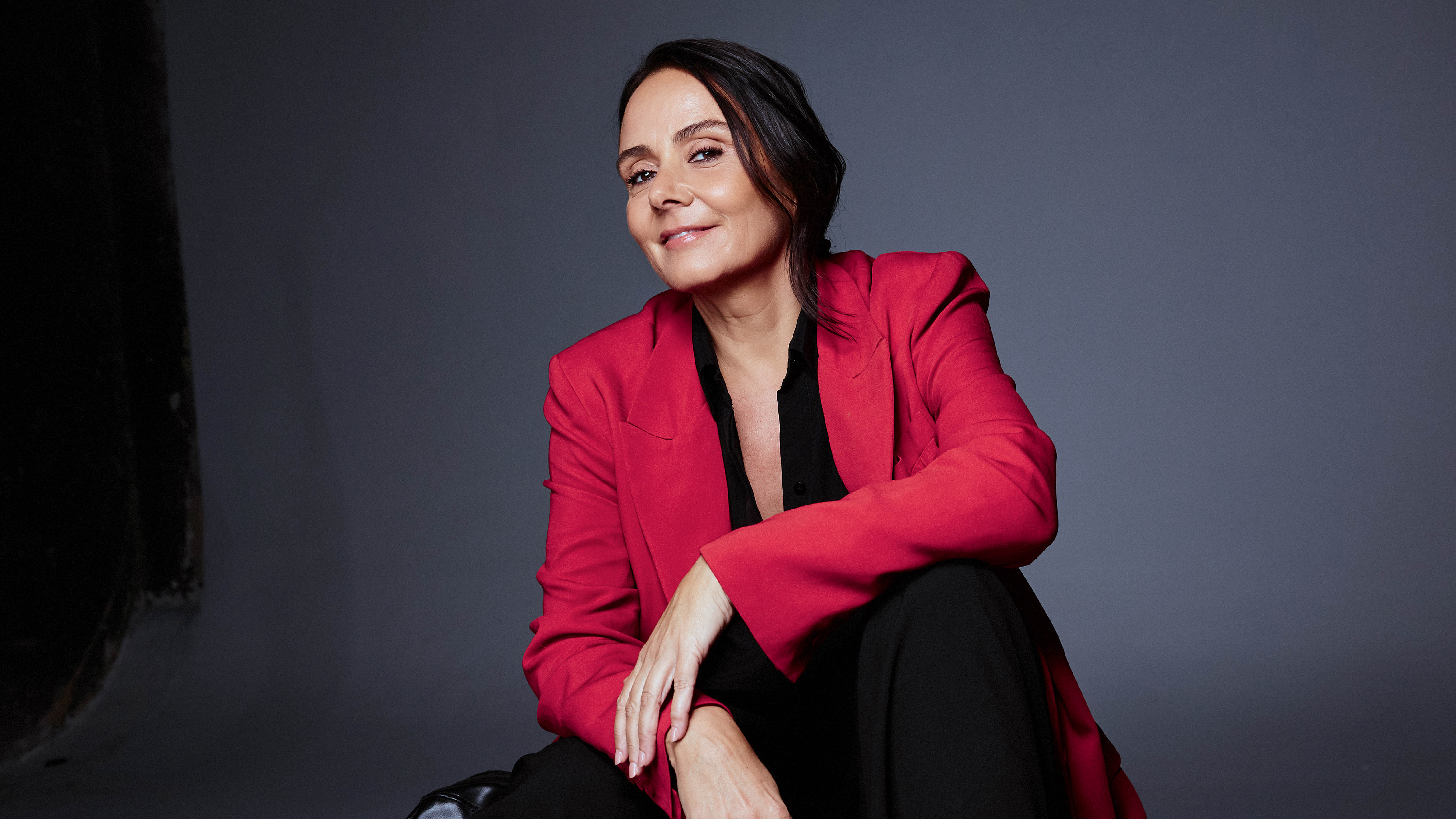 Mulher de meia-idade, cabelos escuros e lisos, sorri levemente para a câmera, vestindo blazer vermelho e blusa preta, sentada em fundo cinza escuro