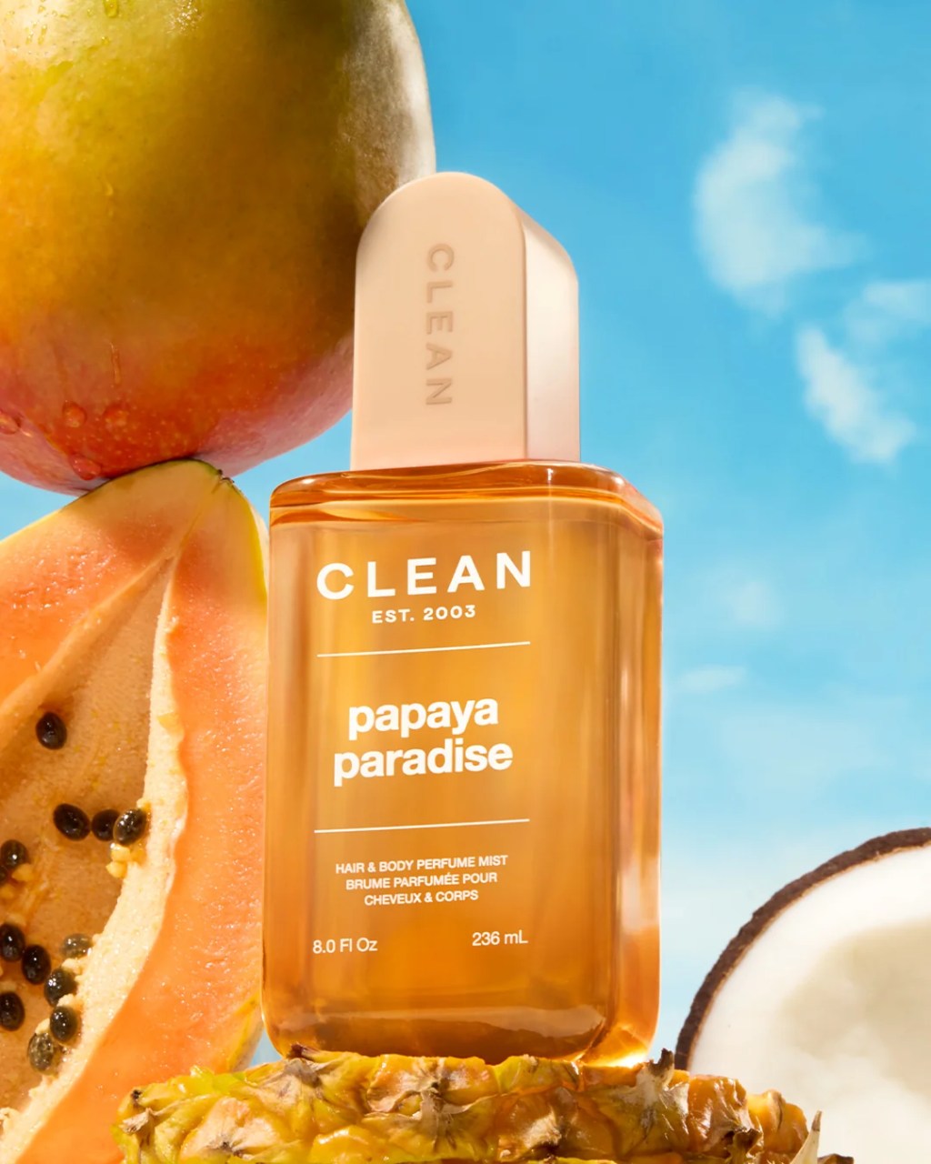 Frasco de perfume Clean Papaya Paradise em tom &acirc;mbar com tampa bege, sobre abacaxi e mam&atilde;o fatiado, com manga e coco ao fundo, sob c&eacute;u azul claro